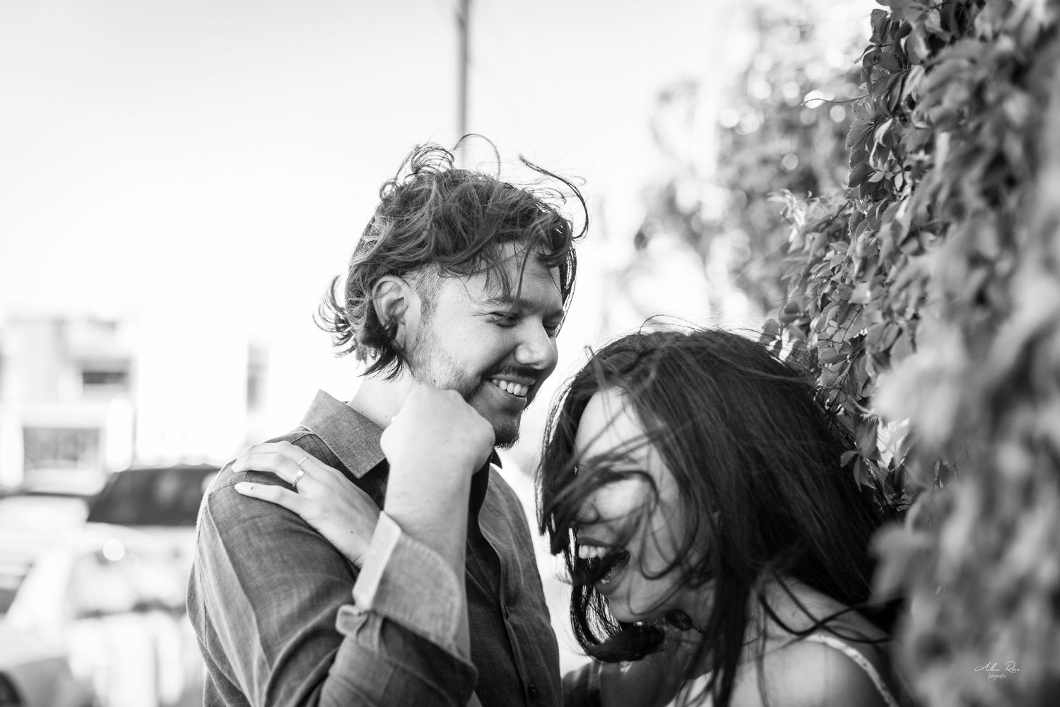 Couple laughs joyfully together outdoors in a candid black and white engagement photo by a leafy wall.