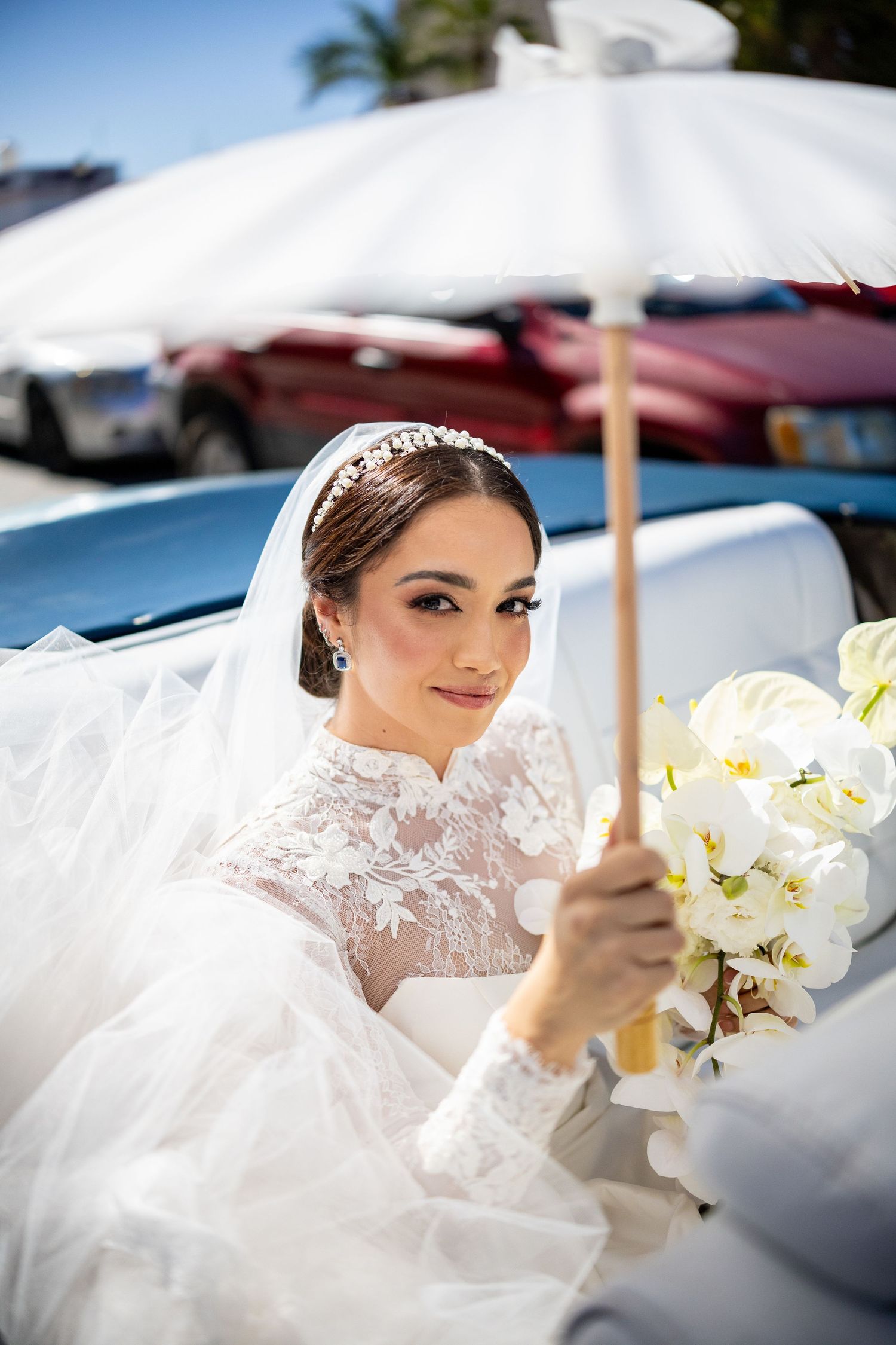 Elegant bride in lace gown holding white parasol and orchid bouquet.
