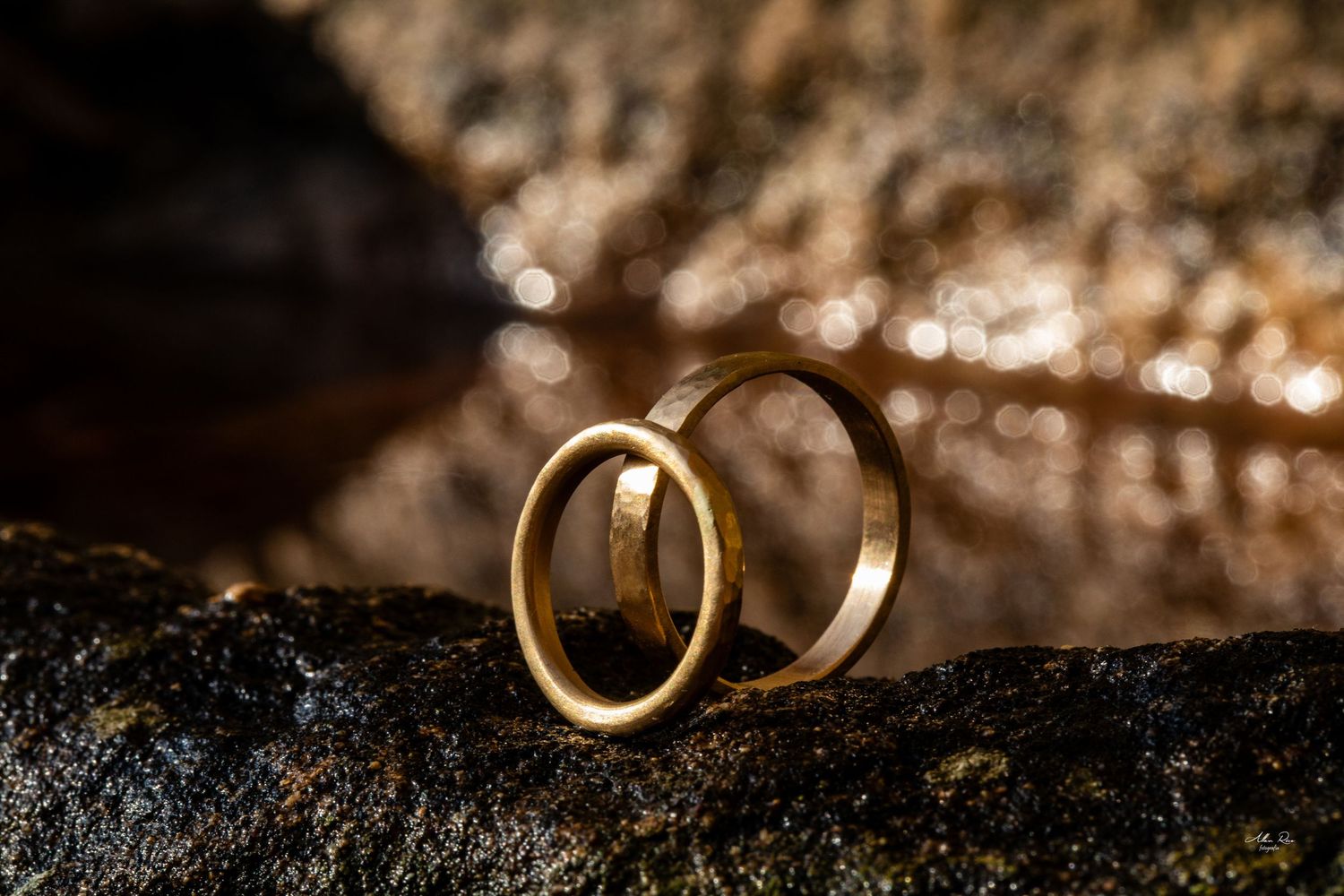 Two gold wedding bands rest together on a dark rocky surface with bokeh background.
