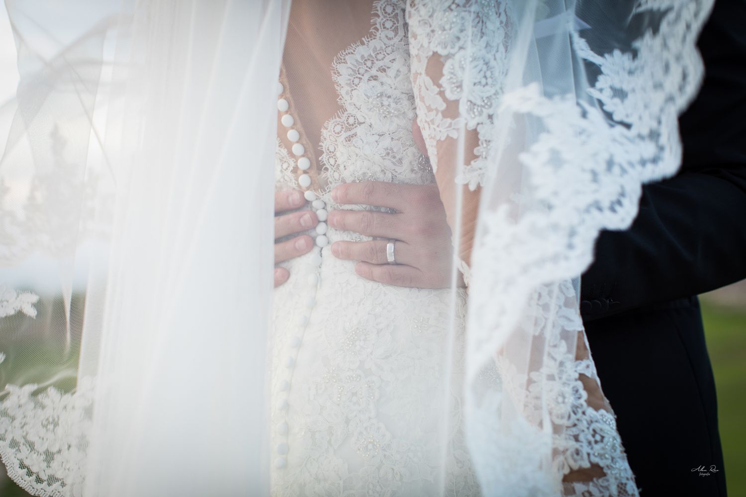 Close-up of brides lace wedding dress and diamond ring visible through delicate lace veil detail.