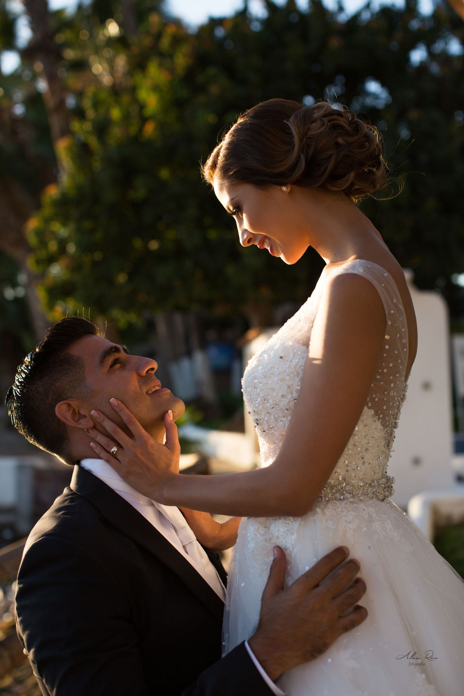 Groom kneels gazing up at bride in beaded gown in a golden sunset portrait.