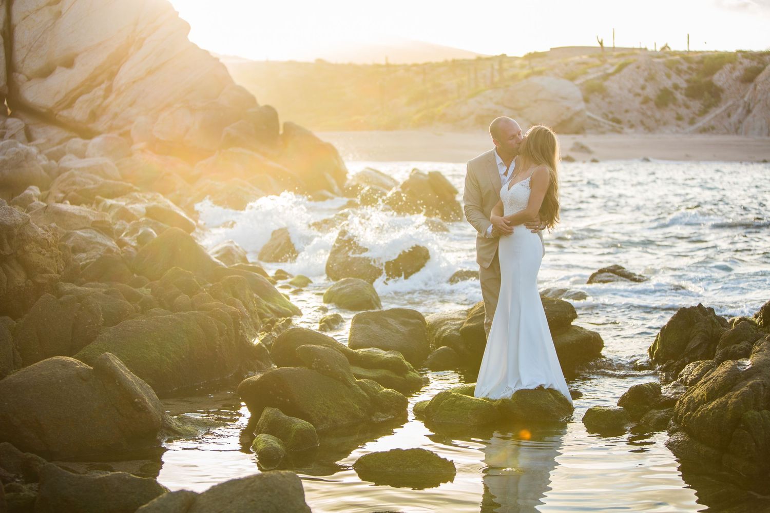 Bride and groom standing on ocean rocks at stunning golden sunset wedding portrait.