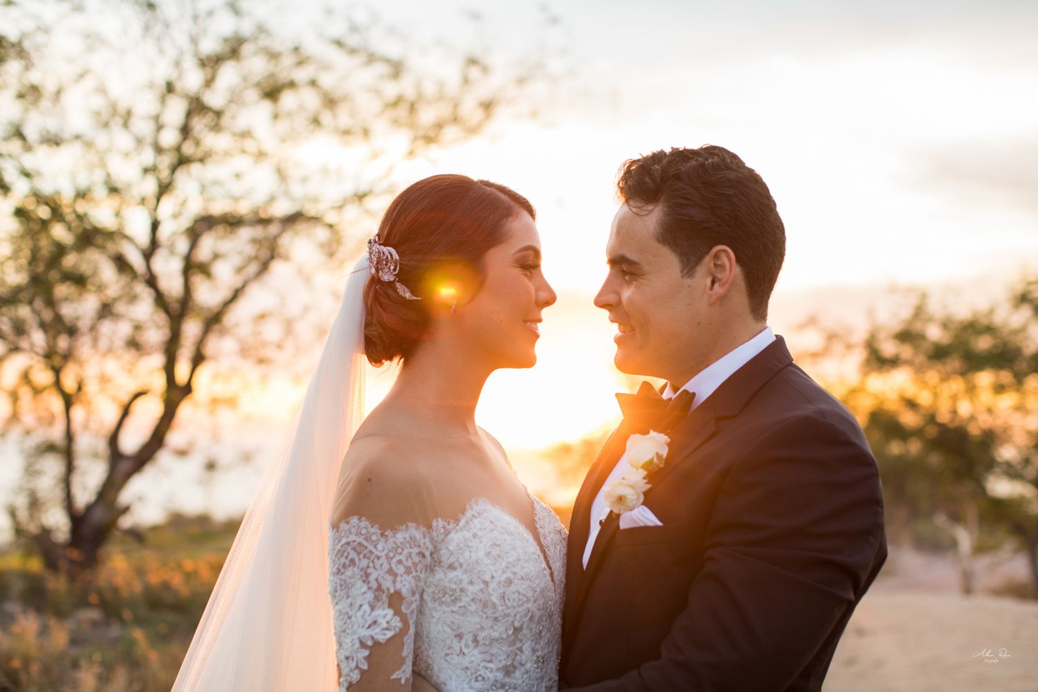 Bride and groom share romantic sunset moment, bride in lace wedding gown with veil, groom in dark suit.
