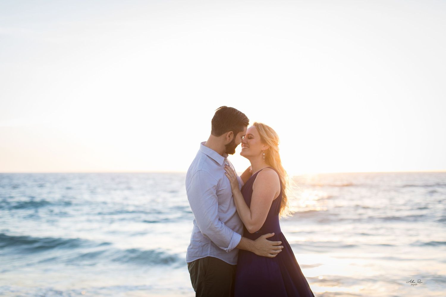 Couple sharing a tender almost-kiss moment at sunset with calm ocean in the background.
