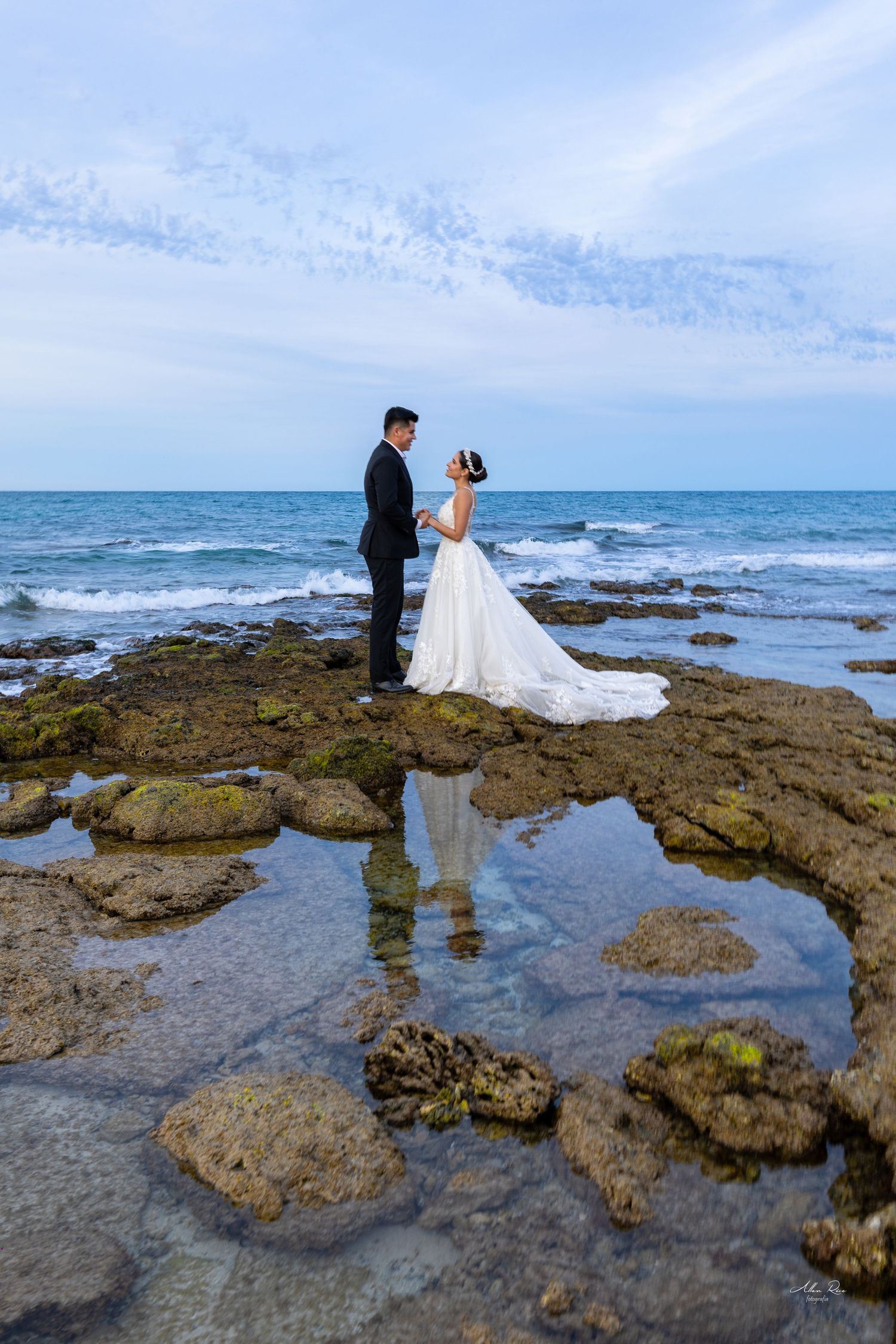 Bride and groom facing each other on rocky coastal shore with ocean behind them.