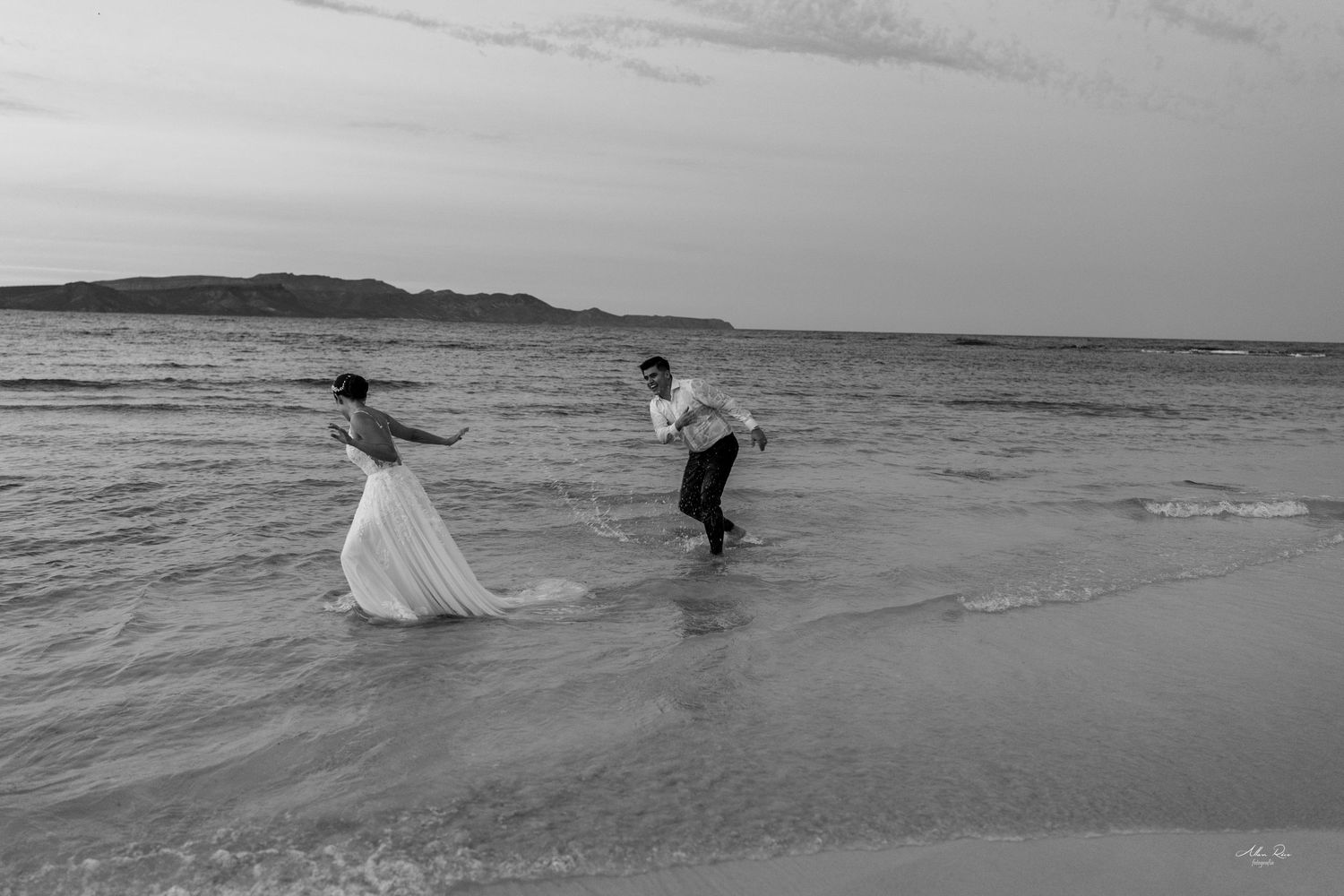 Black and white photo of bride and groom running playfully along shoreline waves at dusk.