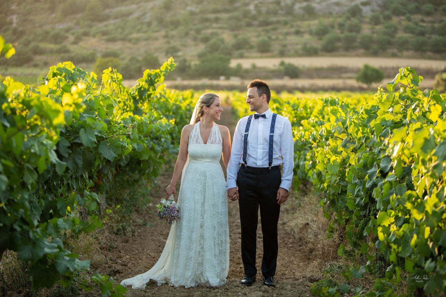 Bride and groom holding hands walking through lush green vineyard rows at golden hour.