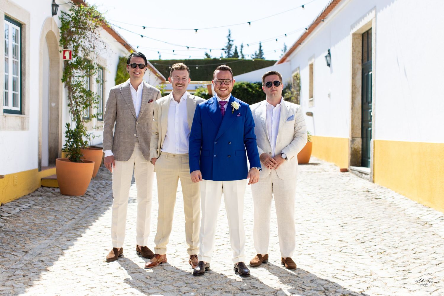Groom in blue suit posing with three groomsmen in tan suits on cobblestone street.