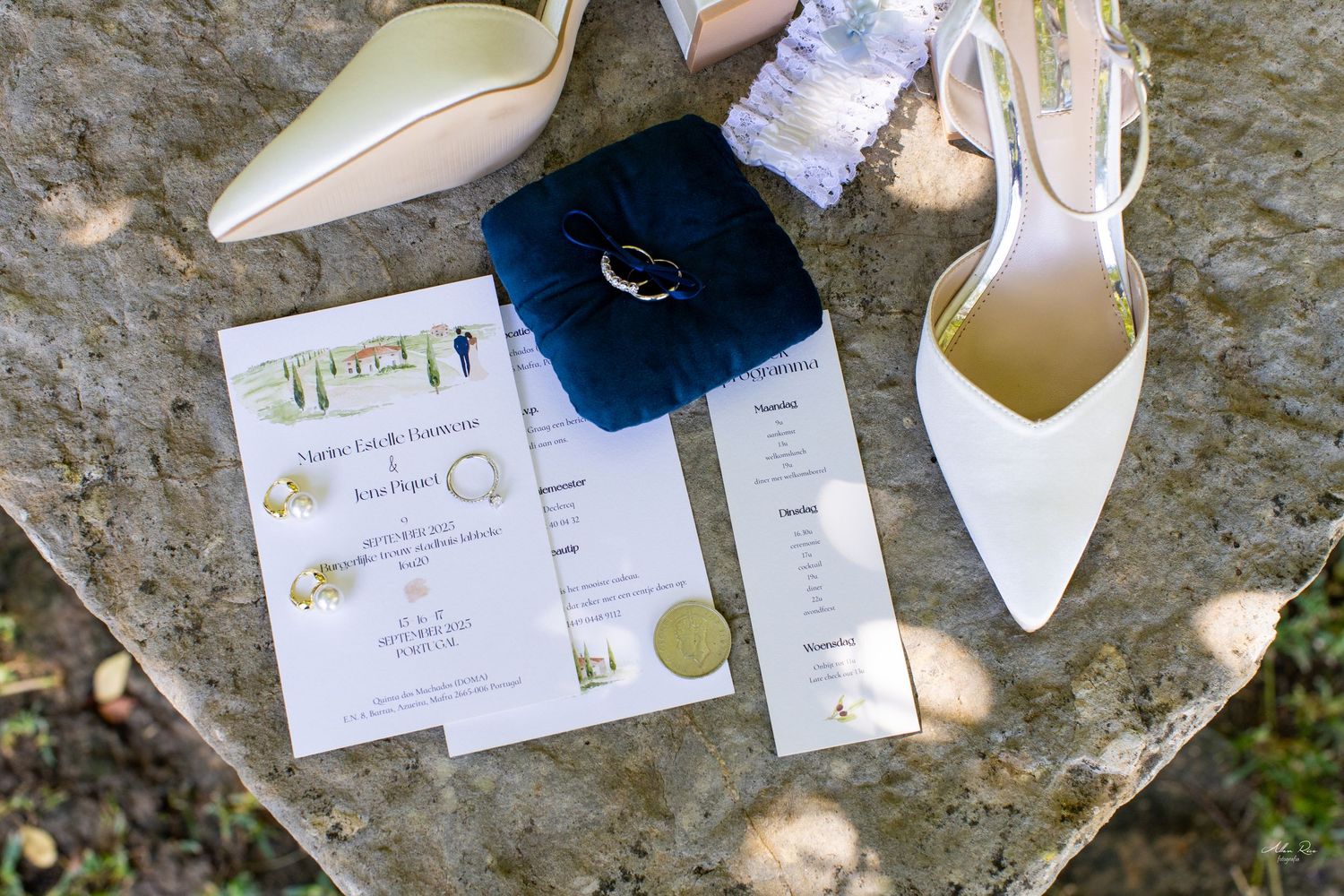 Wedding flat lay with white heels, rings, invitation, and ceremony program on stone.