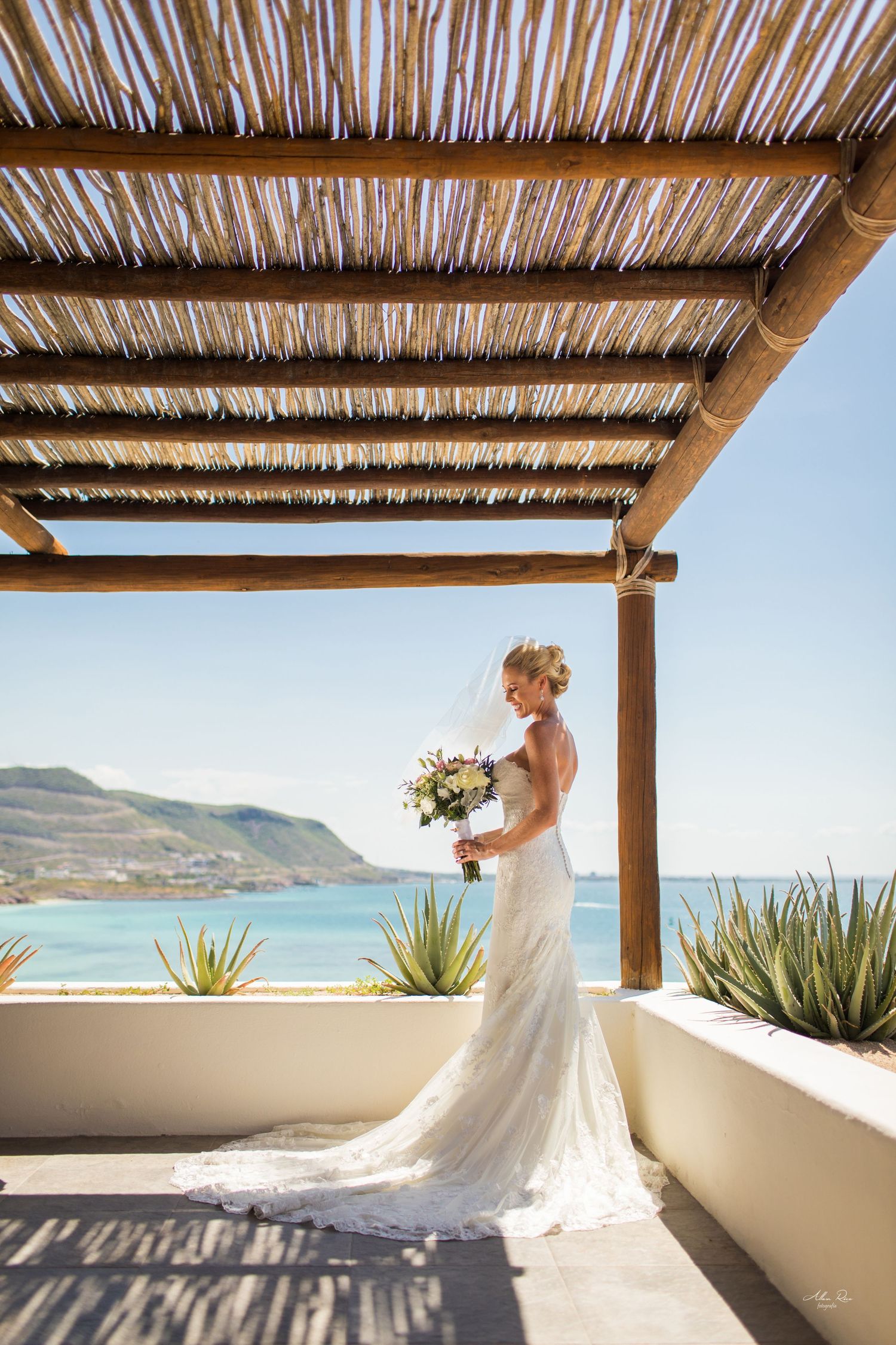 Bride in lace gown holding bouquet under rustic pergola with blue ocean backdrop.