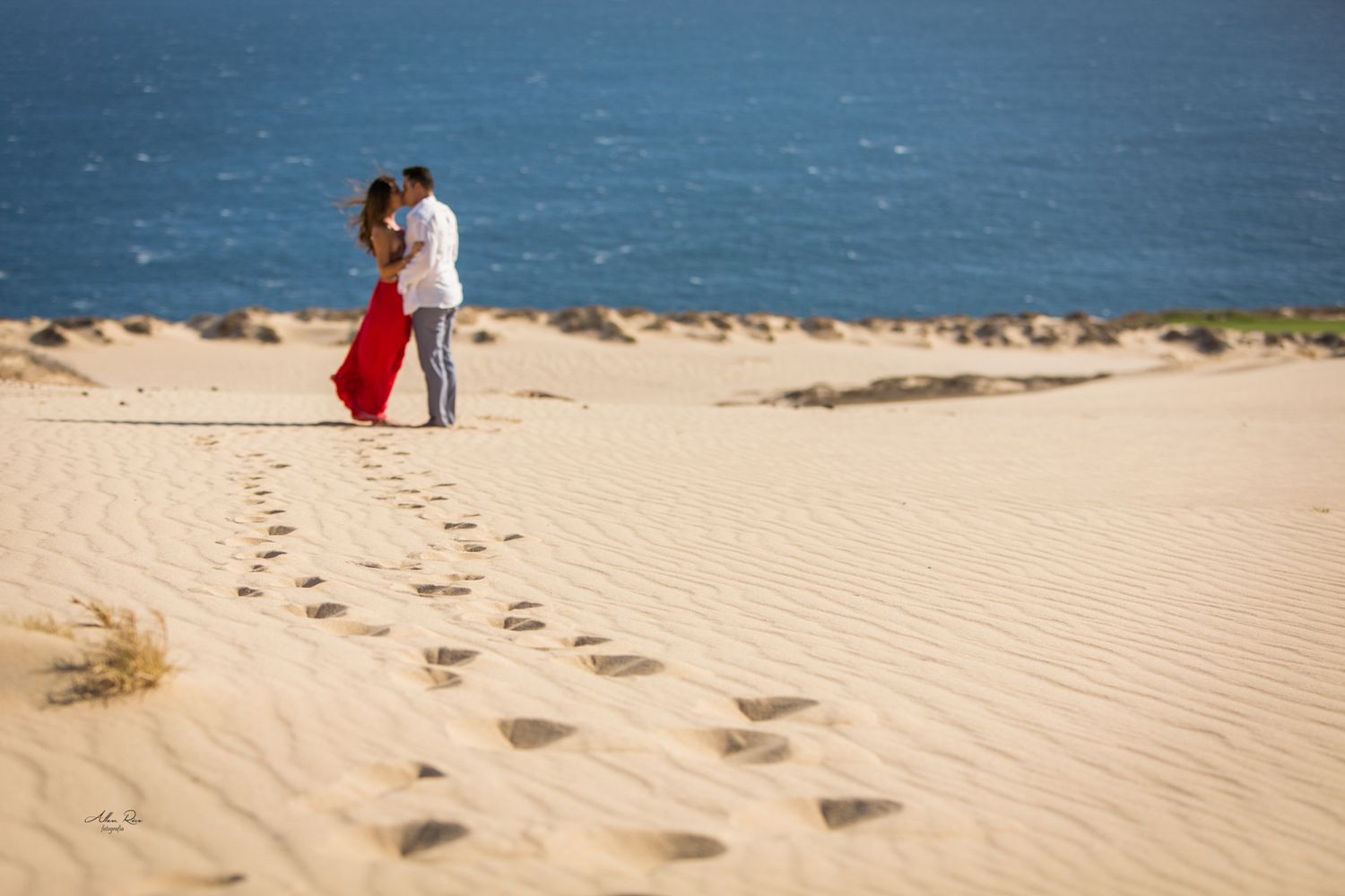Romantic couple kissing on sand dunes with ocean backdrop and footprints in sand.