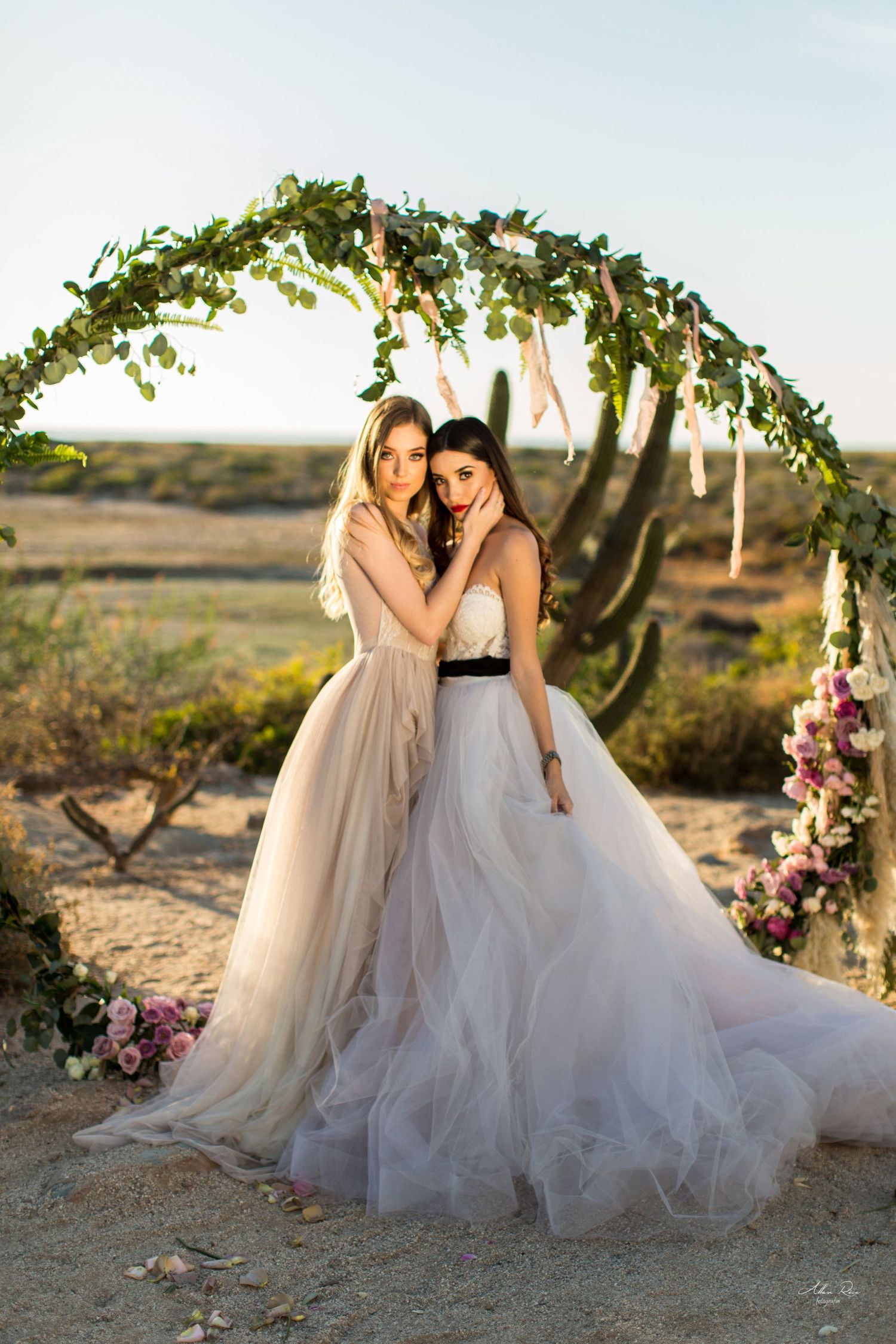 Two brides in elegant tulle wedding dresses pose together under a greenery arch in a desert landscape.