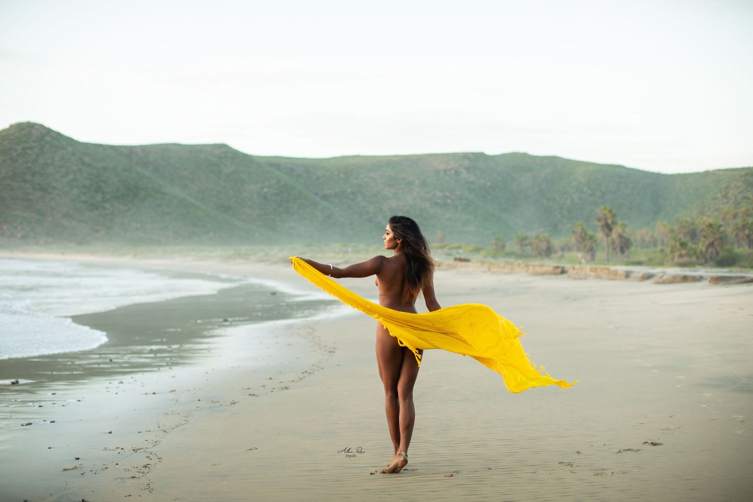 Woman on beach holding flowing yellow fabric, with green hills and ocean in background.