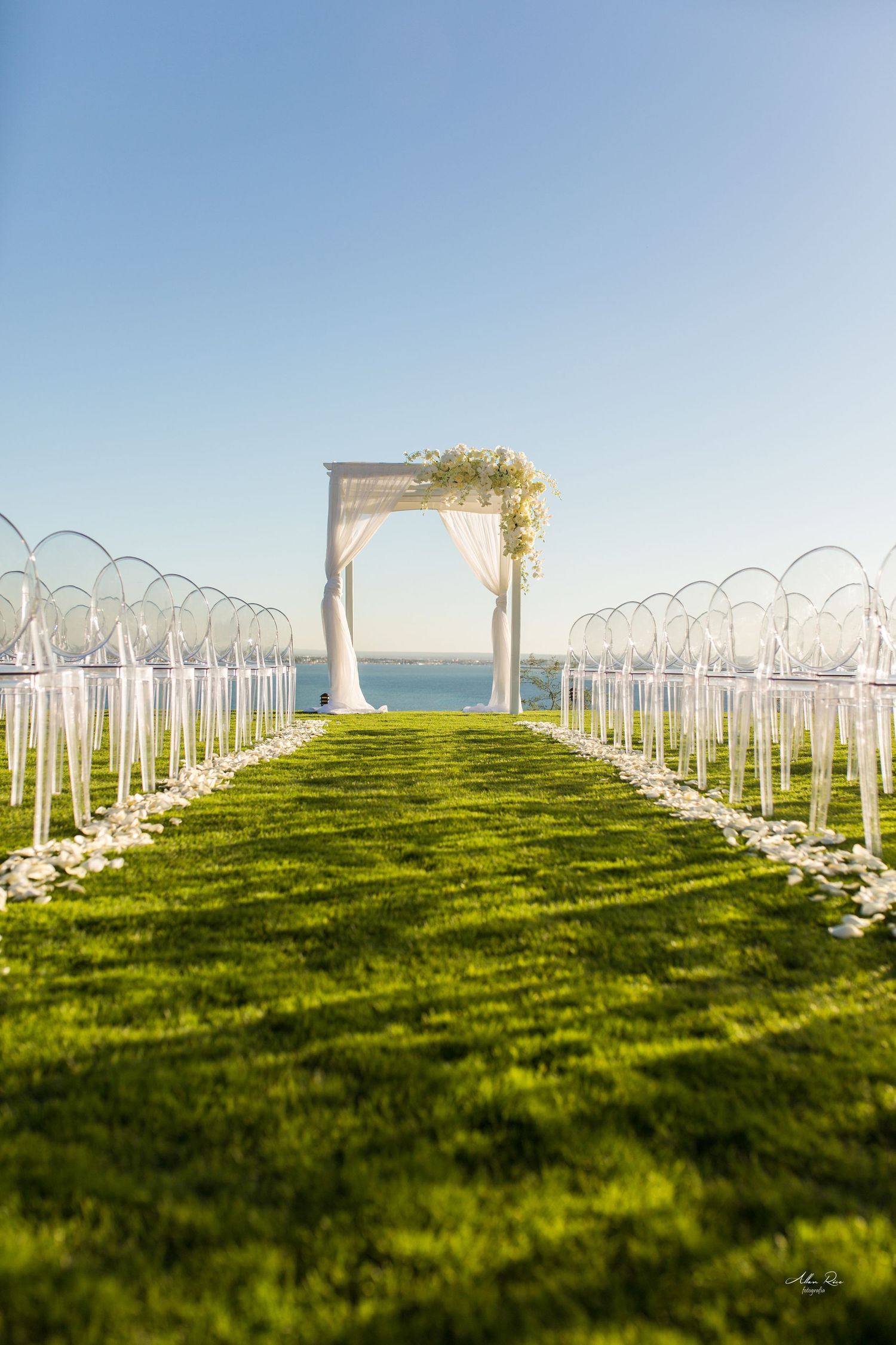 Elegant outdoor wedding aisle with white floral arch, ghost chairs, and rose petals overlooking ocean.