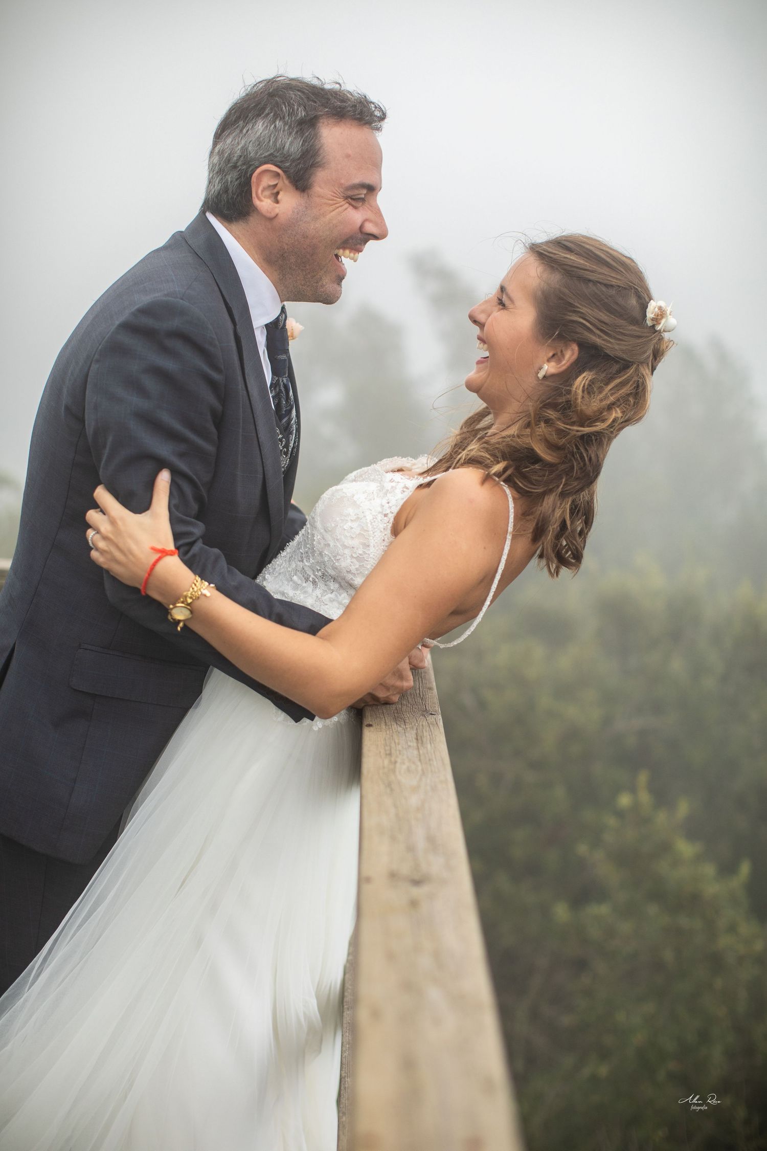 Joyful bride and groom laugh together while embracing on misty wooden railing.