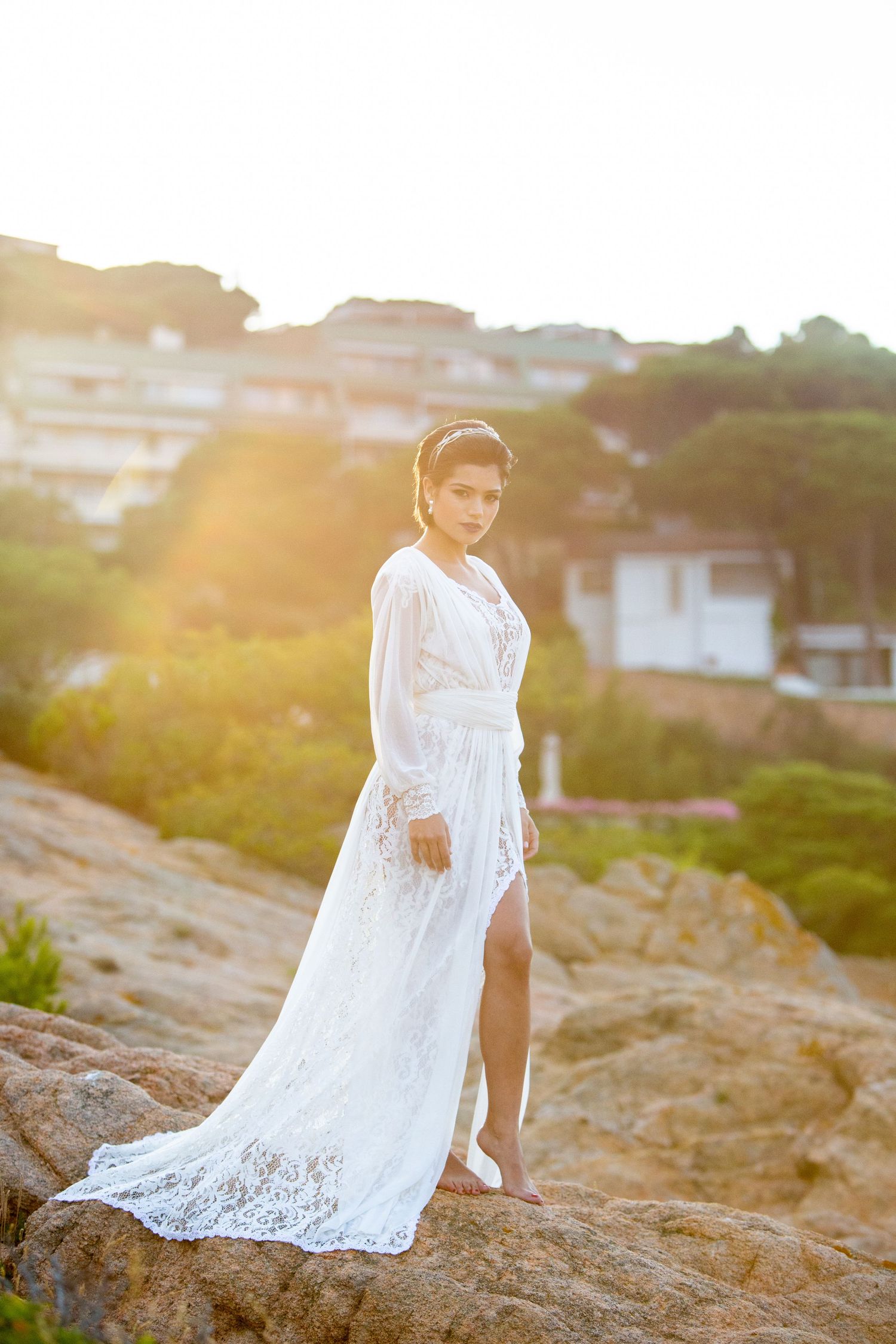 Woman in white lace wedding gown standing on rocks at golden sunset with coastal village in background.