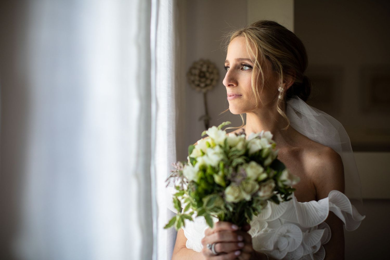 Elegant blonde bride in white gown gazing out a window while holding a bouquet of white and green flowers.
