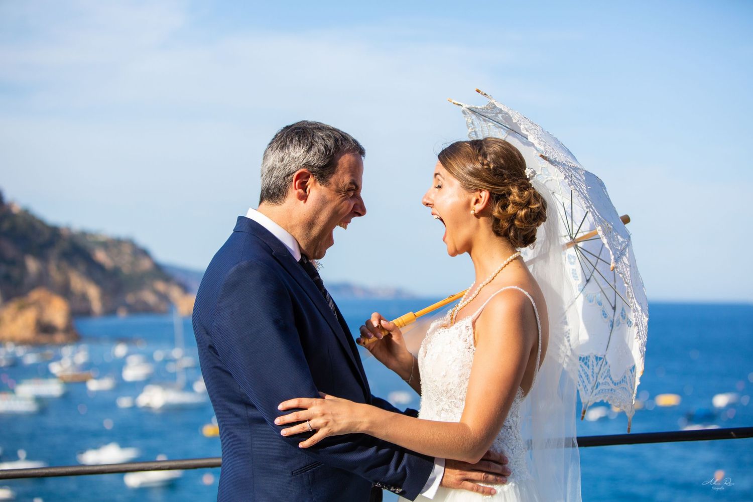 Bride laughing joyfully with groom while holding a lace parasol against a stunning blue ocean backdrop.