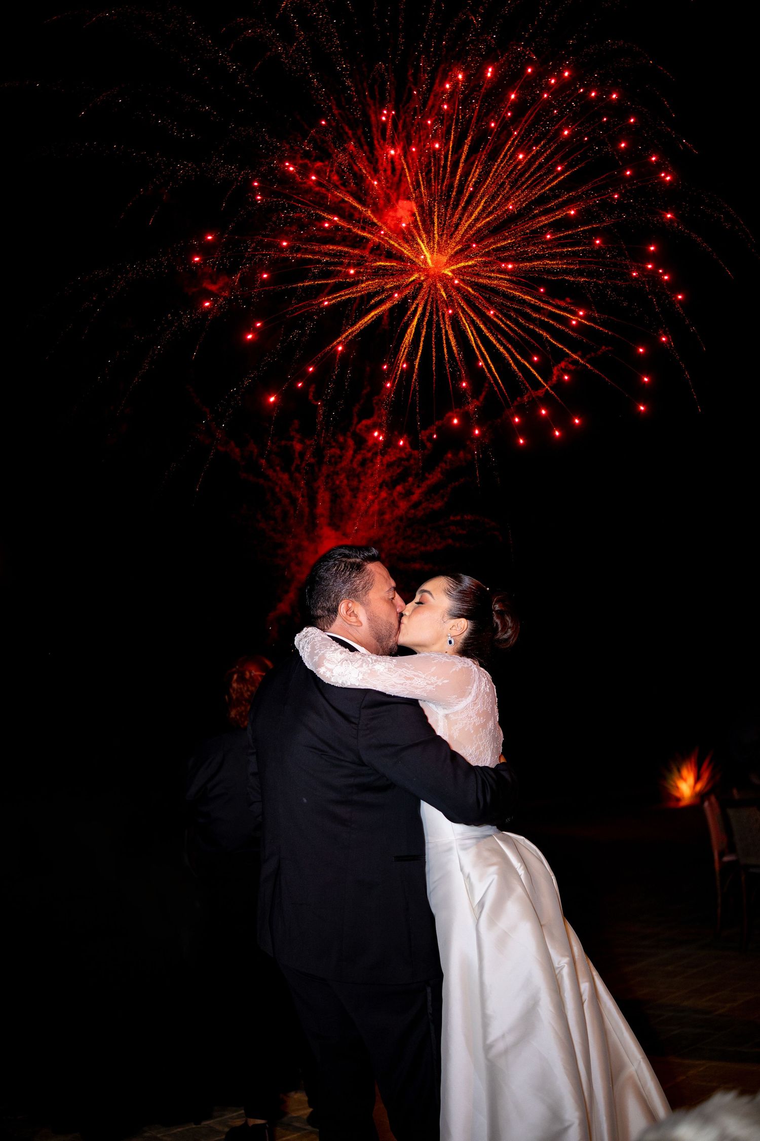 Bride and groom share a romantic kiss beneath stunning red fireworks display on their wedding night.