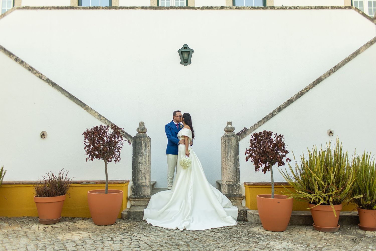Wedding couple embraces before white estate wall with yellow potted plants.