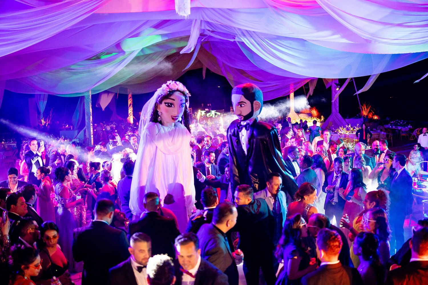 Giant bride and groom puppet figures tower over guests on a vibrant, purple-lit wedding dance floor.