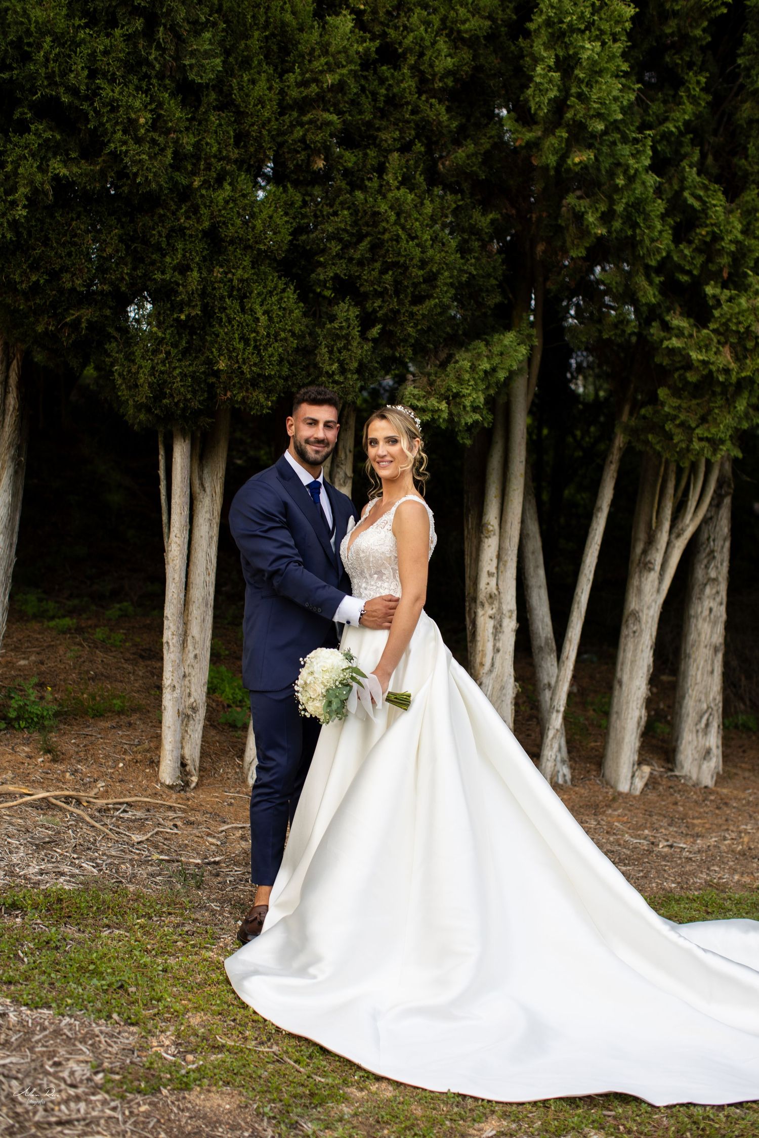 Elegant bride in white ball gown holds bouquet as groom embraces her among tall cypress trees.