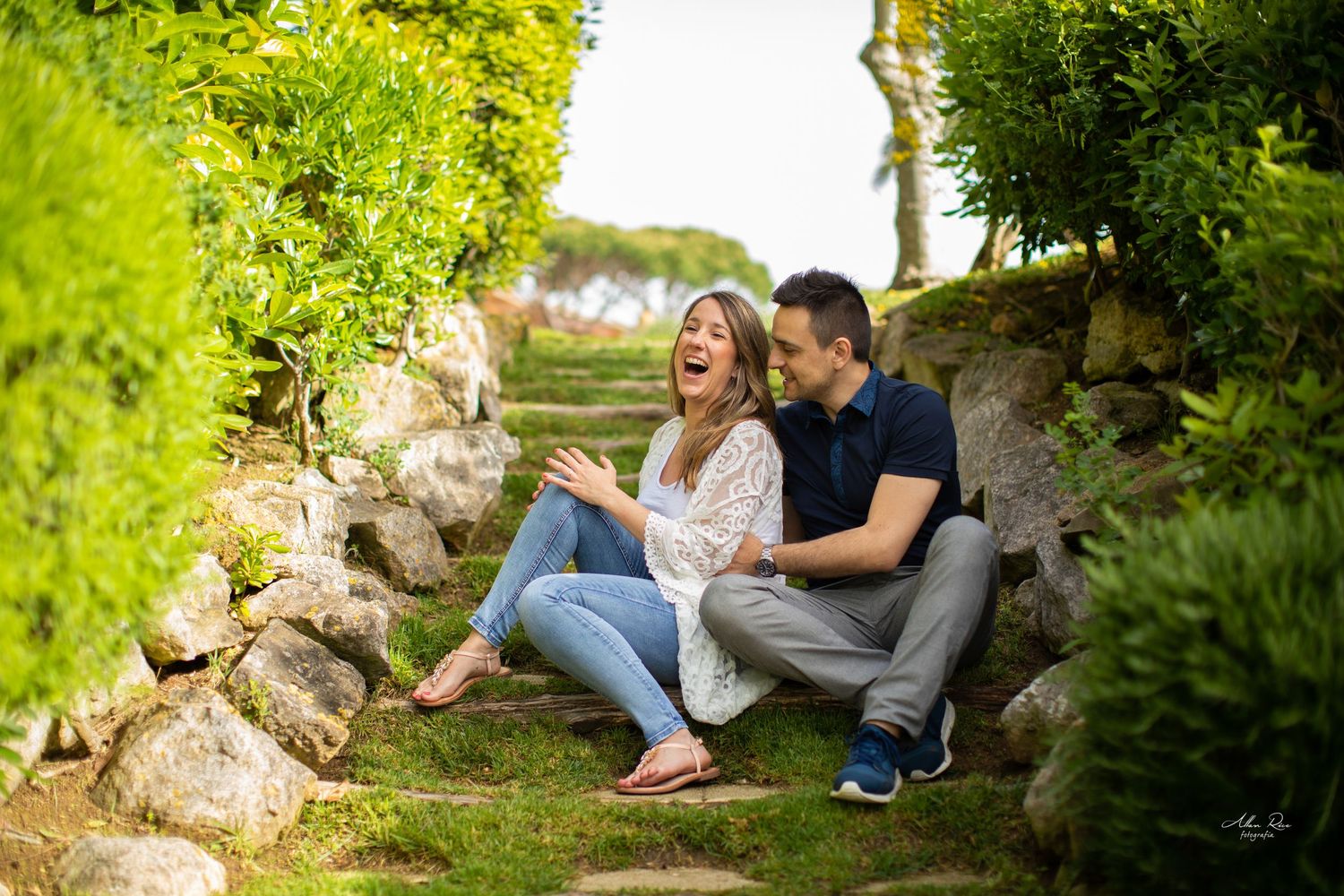 Couple laughing joyfully while sitting on stone steps surrounded by lush green gardens.