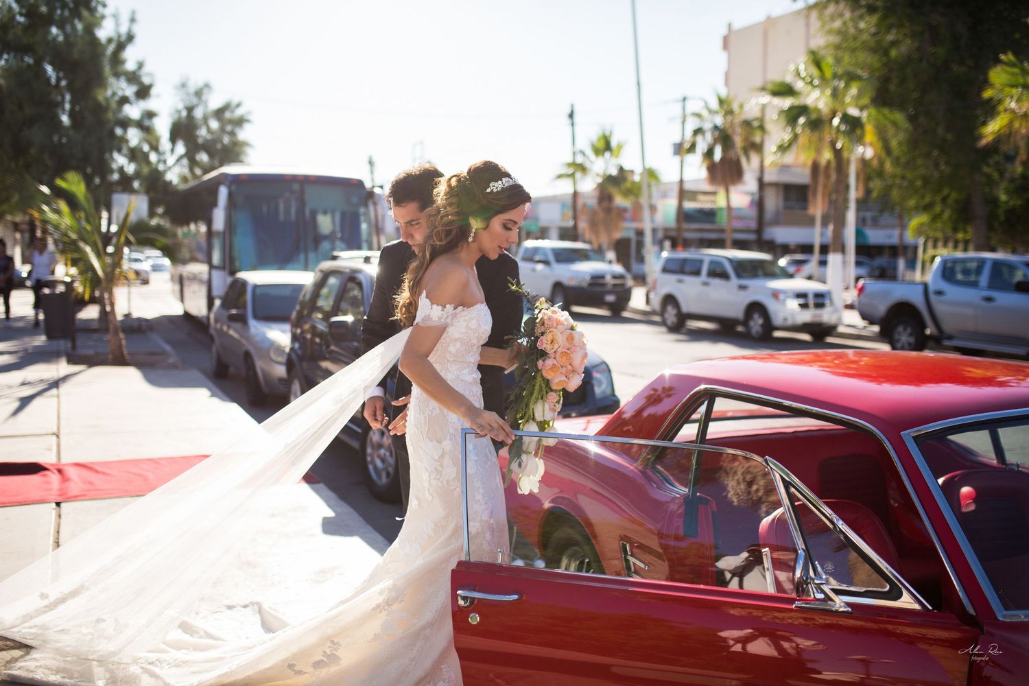 Bride in off-shoulder gown with long veil stepping into red classic car on sunny city street.