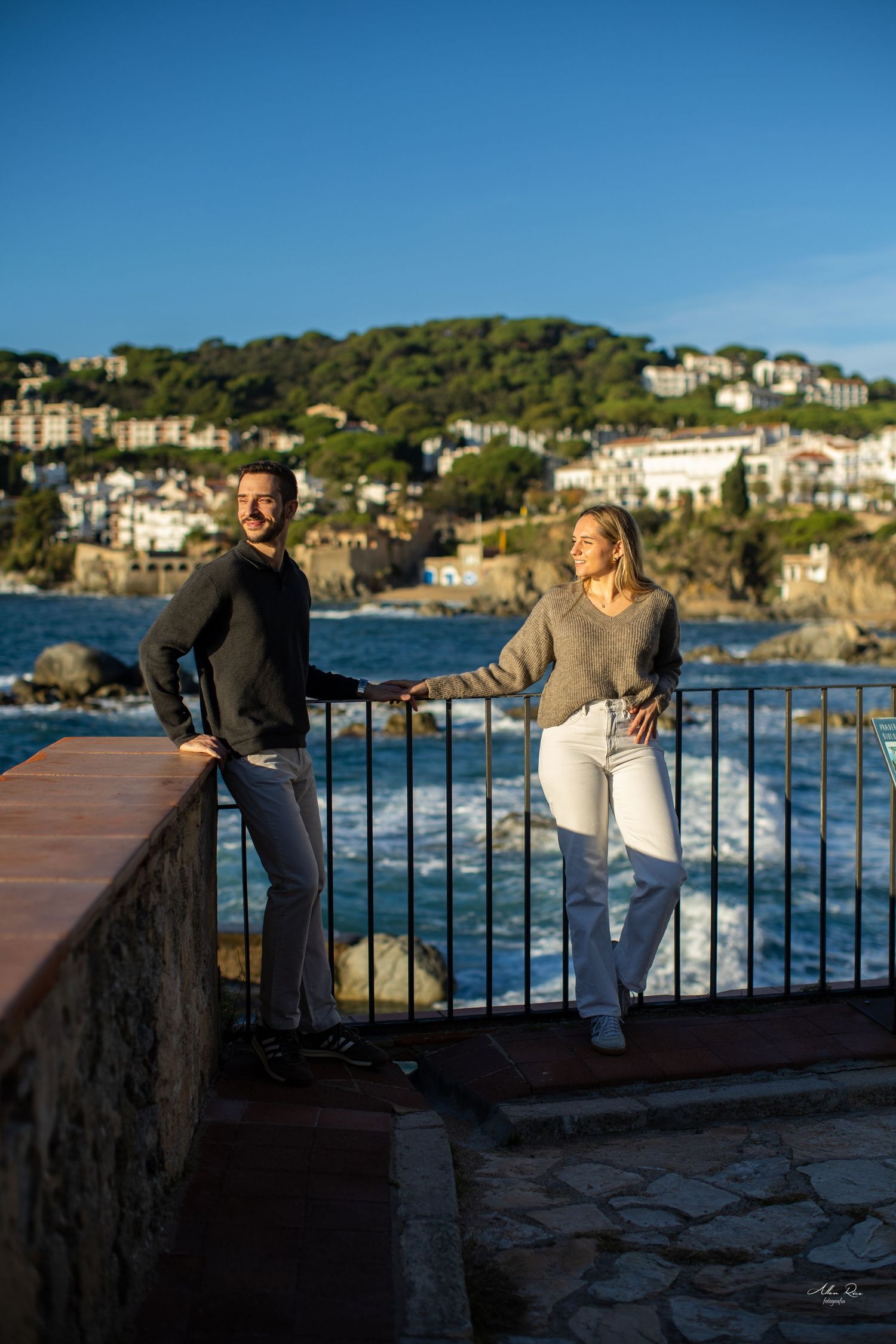 Couple holding hands by coastal railing with rocky shoreline and hillside village in background on sunny day.