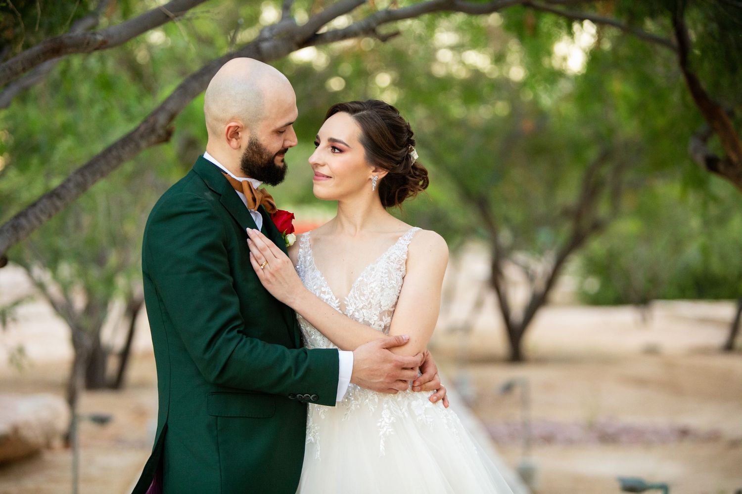 Bride and groom share a romantic embrace outdoors among green trees on their wedding day.