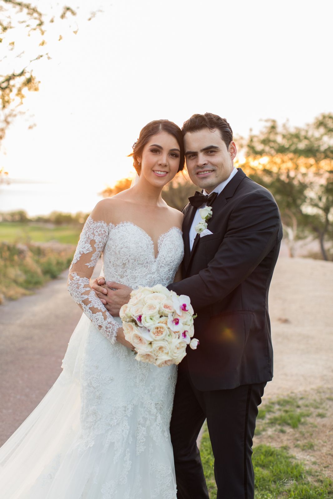 Bride and groom pose at sunset, bride holding white floral bouquet in elegant lace wedding dress.