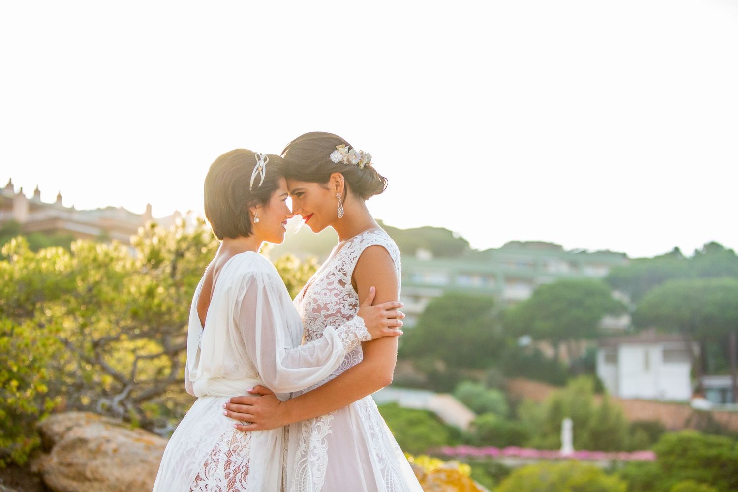 Two brides in white lace wedding dresses embrace tenderly at golden hour with scenic hillside backdrop.