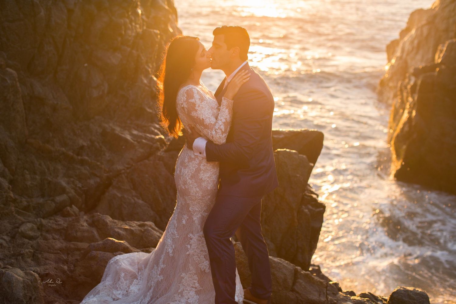 Bride and groom sharing romantic kiss on rocky coastline bathed in warm golden sunset light.