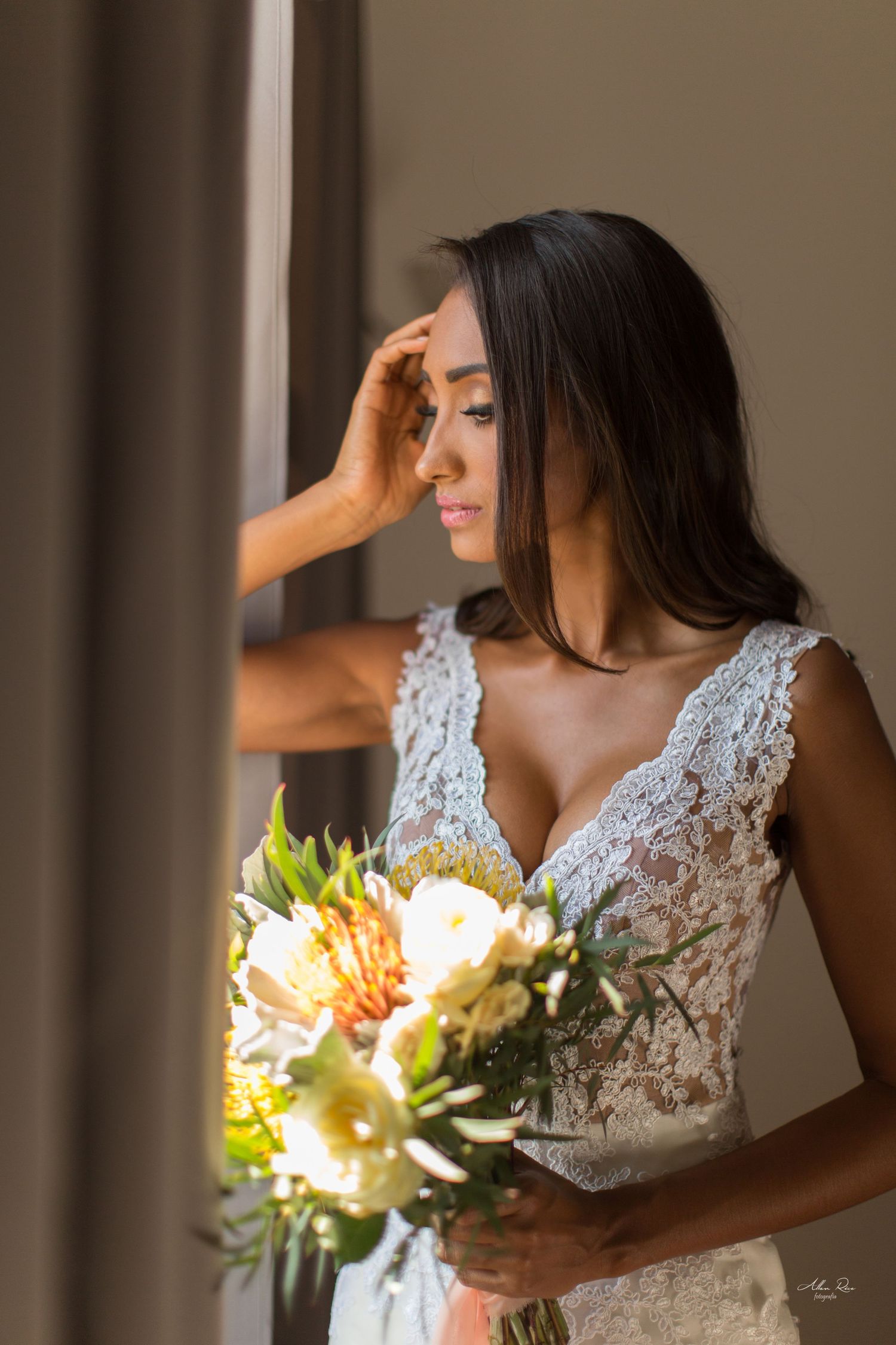 Elegant bride in lace gown holds colorful bouquet while gazing pensively out a sunlit window.
