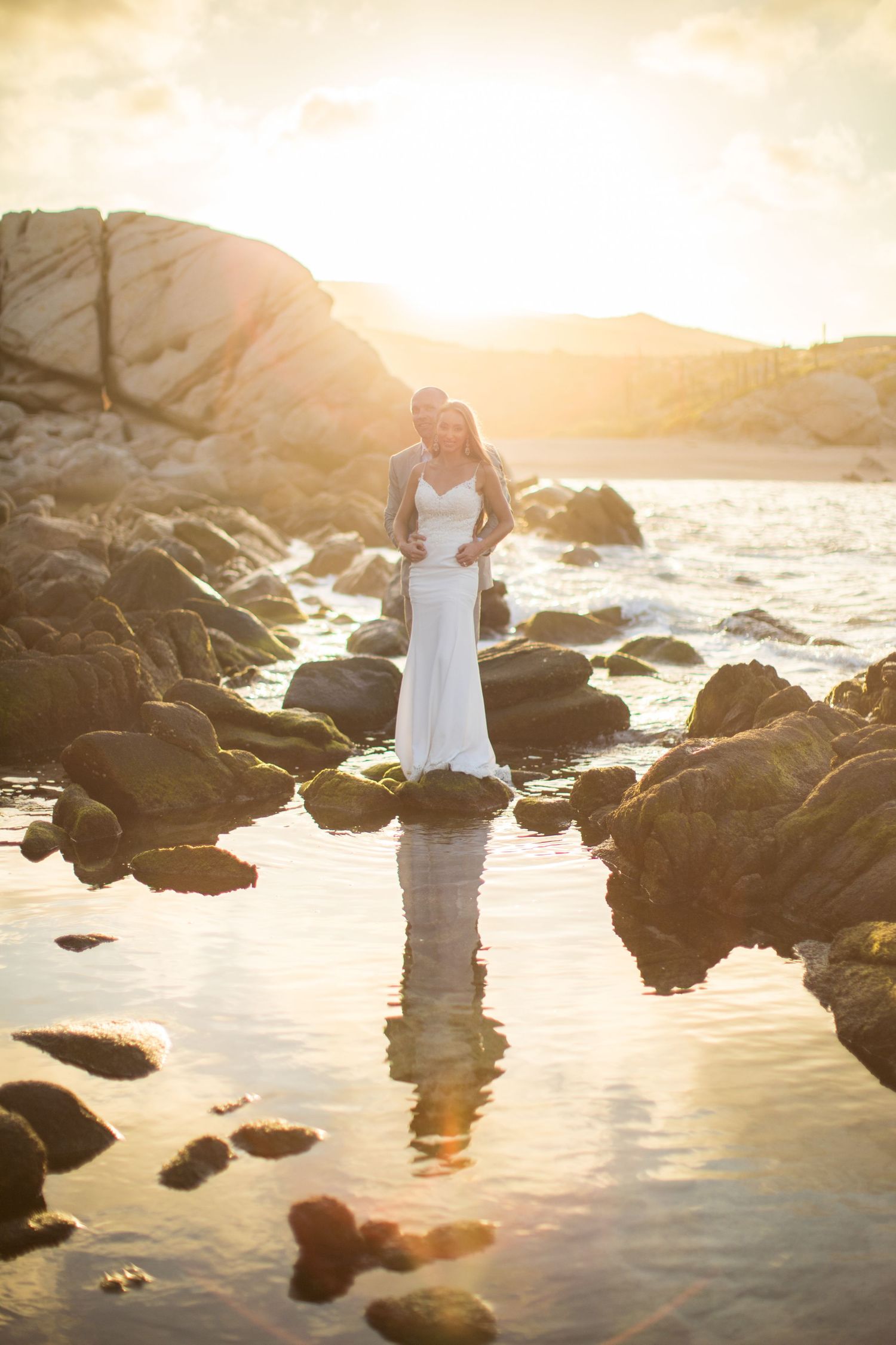 Couple in wedding attire stand on rocks reflected in tidal pools during a golden sunset.