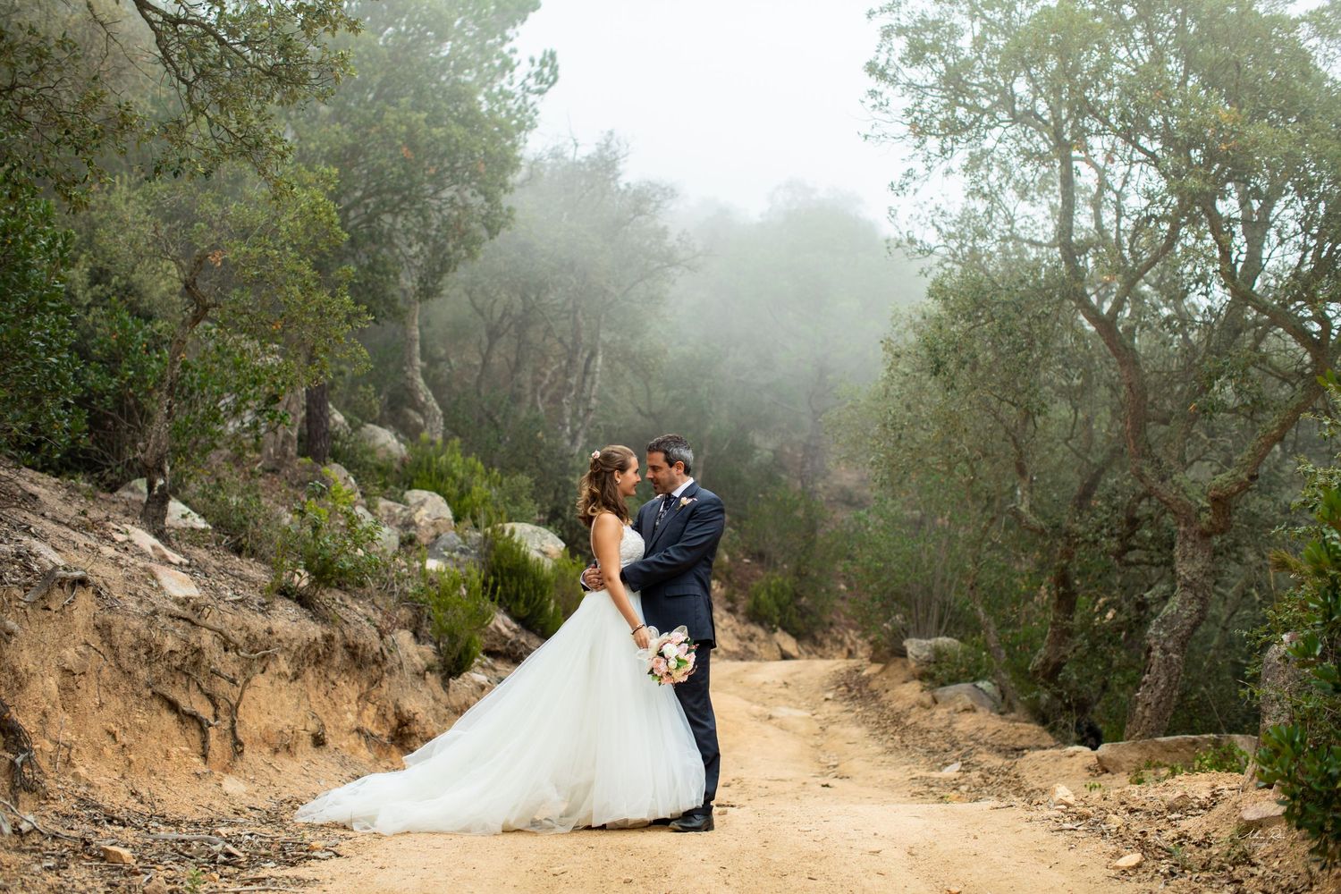 Bride and groom embrace on misty forest dirt path surrounded by trees.