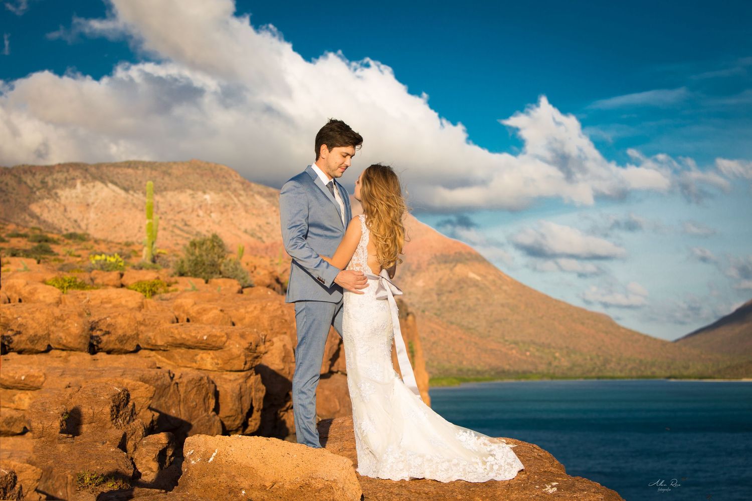 Bride and groom holding hands on red rocks with stunning mountain lake backdrop under blue sky.