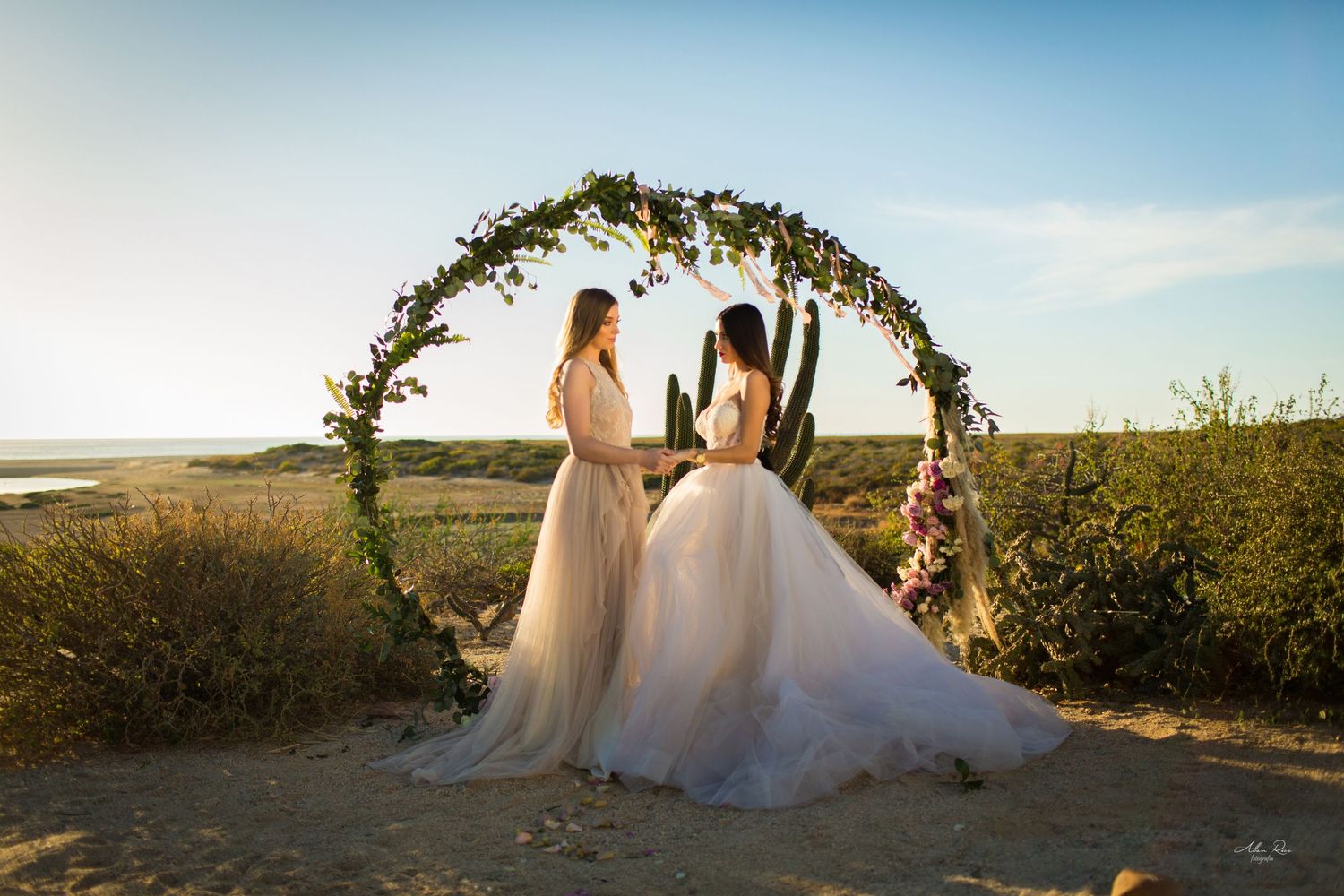 Two brides in white gowns exchange vows under a circular floral arch in a desert landscape.