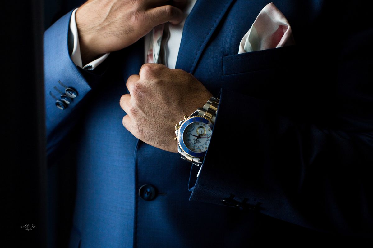 Groom in blue double-breasted suit adjusting pocket square, showcasing luxury gold watch on wrist.