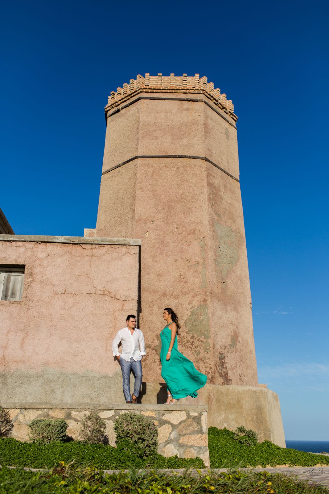 Couple posing beside ancient stone watchtower with woman in turquoise dress against bright blue sky.