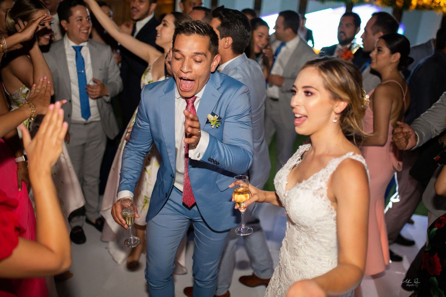 Groom in light blue suit dancing energetically with bride at lively wedding reception party.