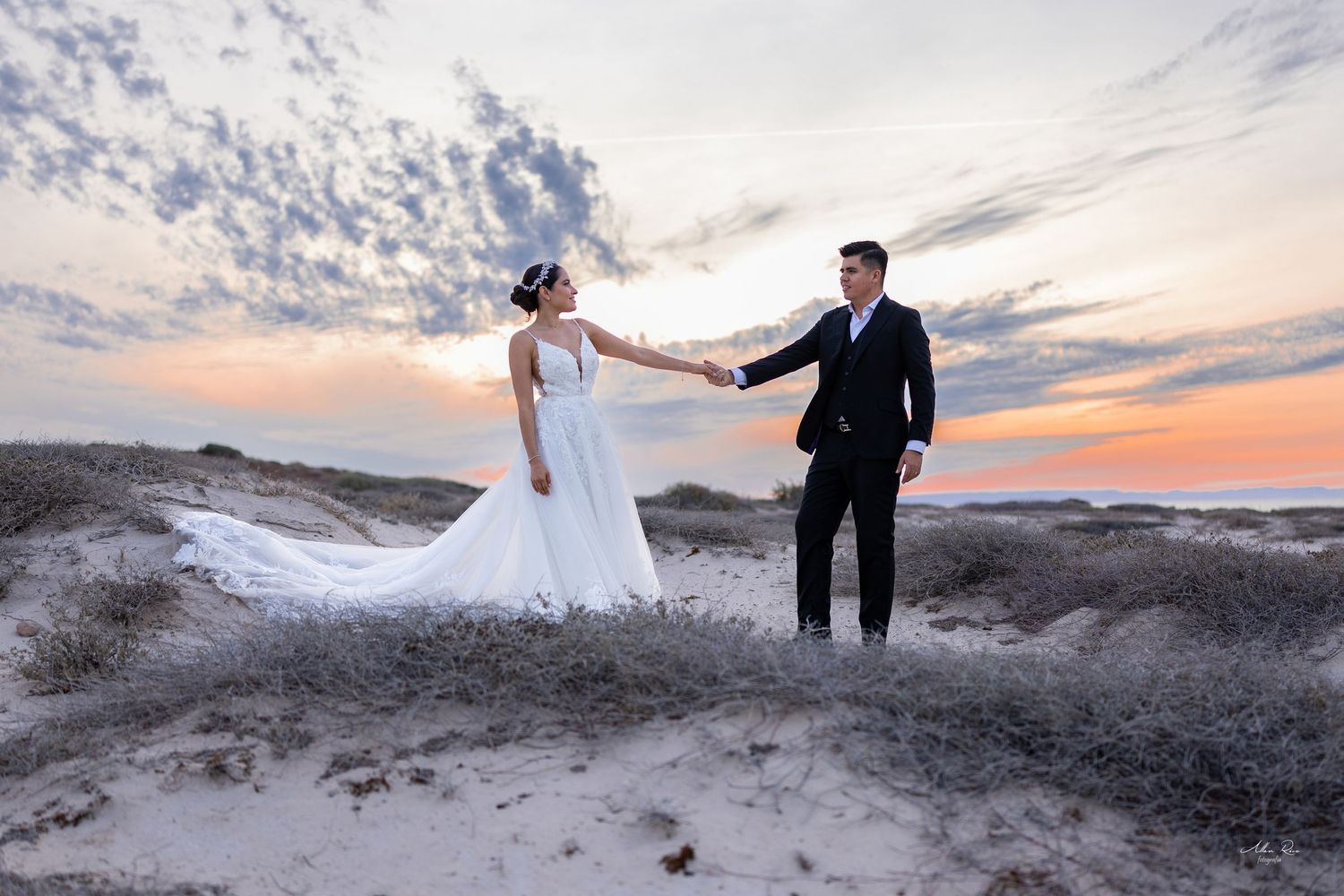 Bride and groom holding hands on sandy dunes at sunset in wedding attire.