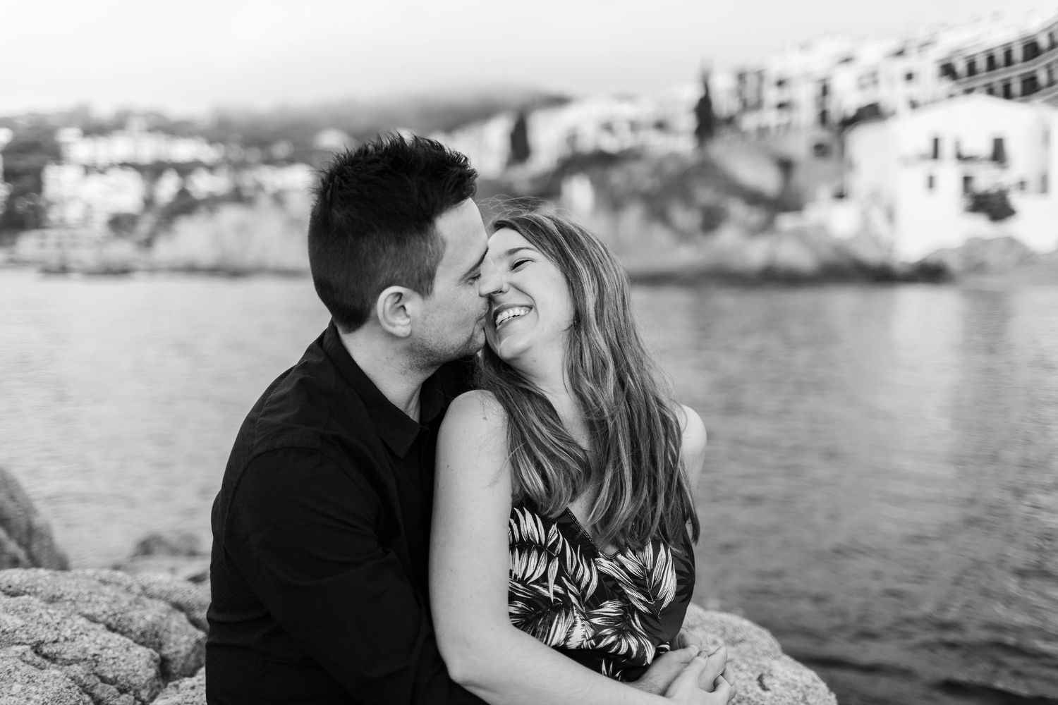 Couple sharing a joyful intimate moment on seaside rocks with a picturesque coastal village behind them.