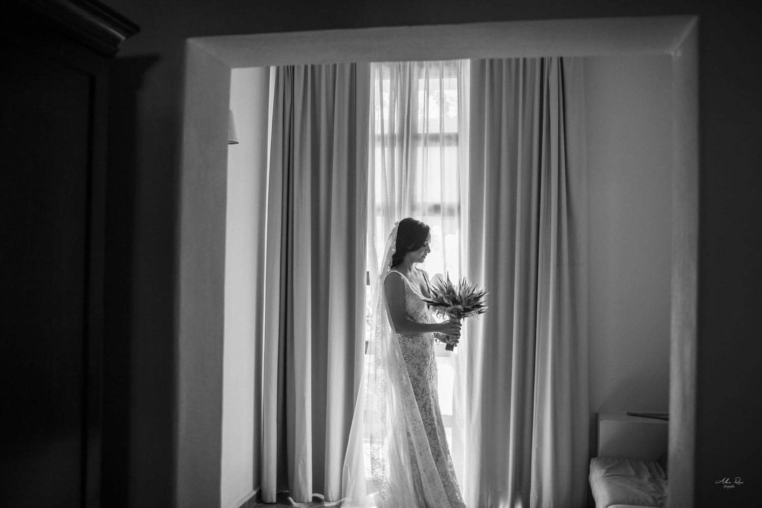 Bride in wedding gown holding bouquet silhouetted against bright window curtains in black and white photo.
