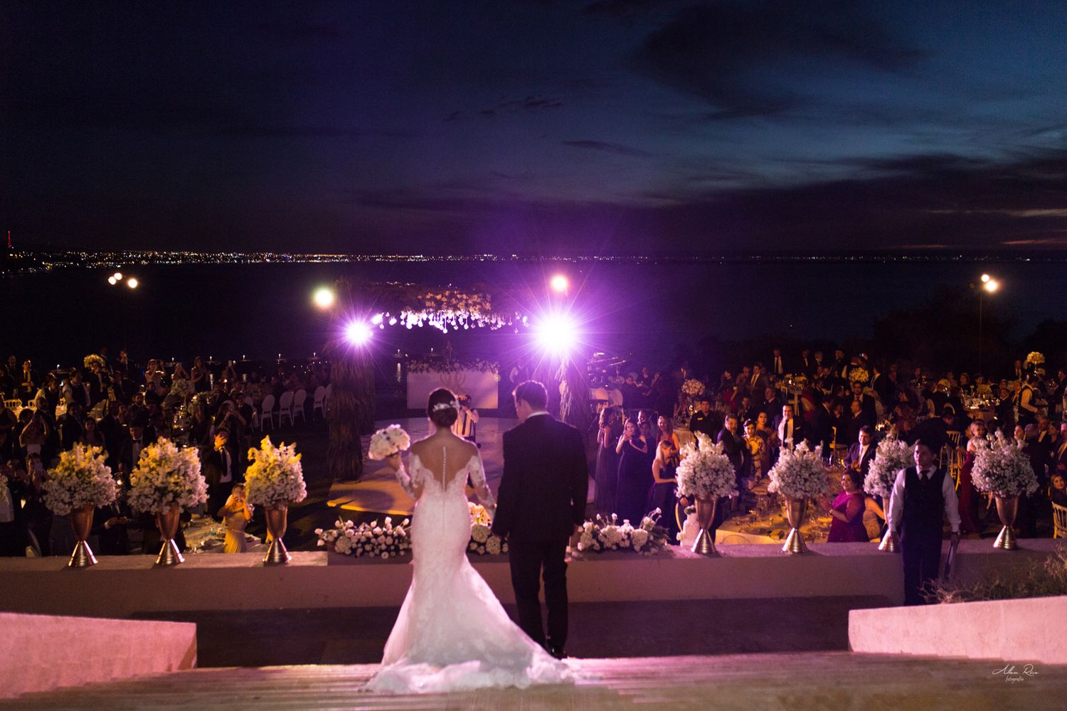 Bride and groom stand before large seated wedding reception crowd at stunning oceanview outdoor venue at night.