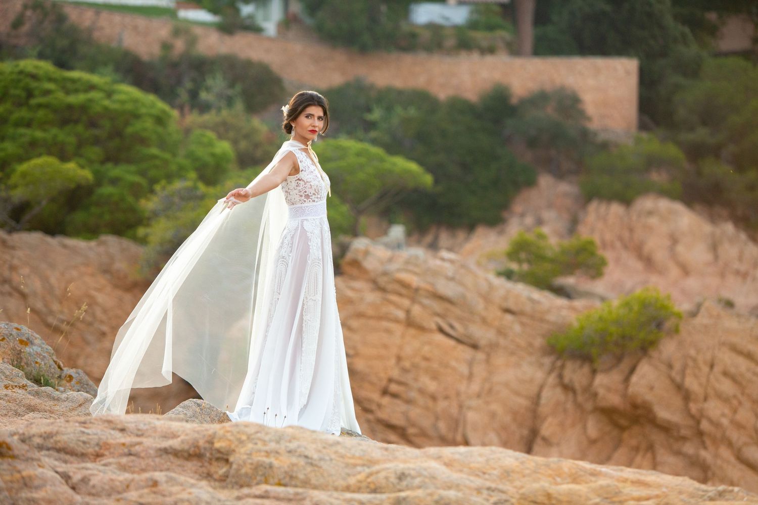 Elegant bride in flowing white lace wedding gown with cape poses gracefully on coastal rocky cliffs.