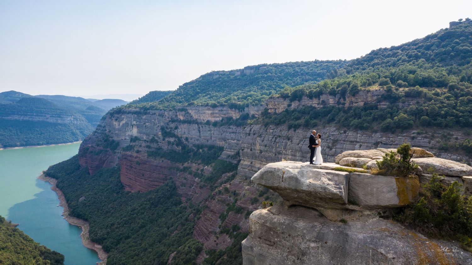 Bride and groom standing on dramatic cliff edge overlooking a stunning turquoise river canyon landscape.