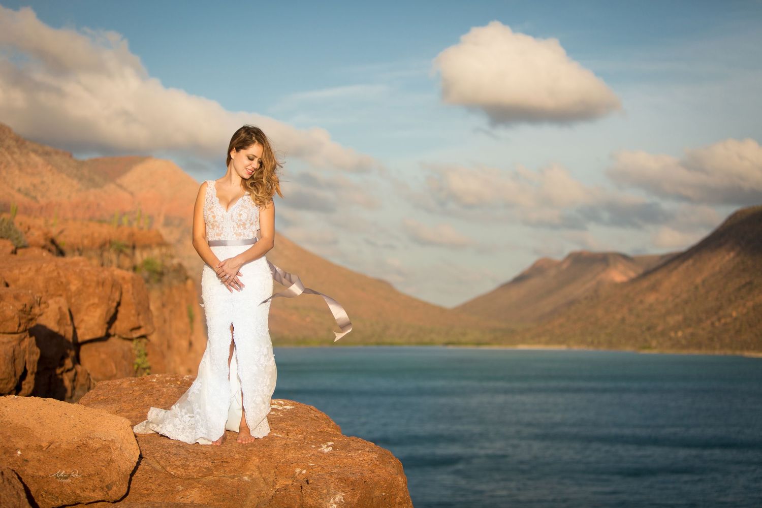 Bride in white lace gown stands on red rock cliff with turquoise lake and mountains at golden sunset.