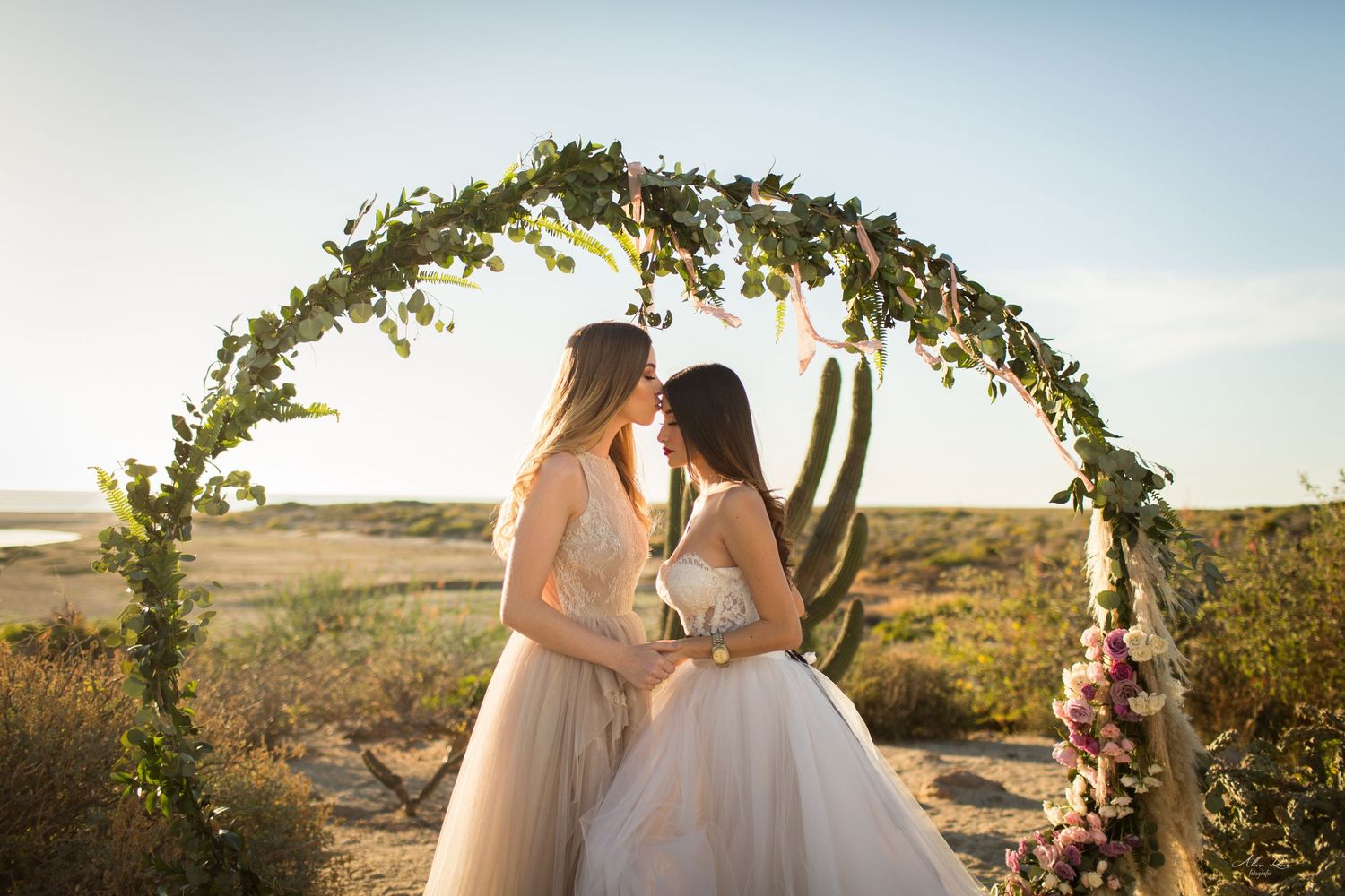 Two brides sharing an intimate moment under a green circular arch in a desert setting.