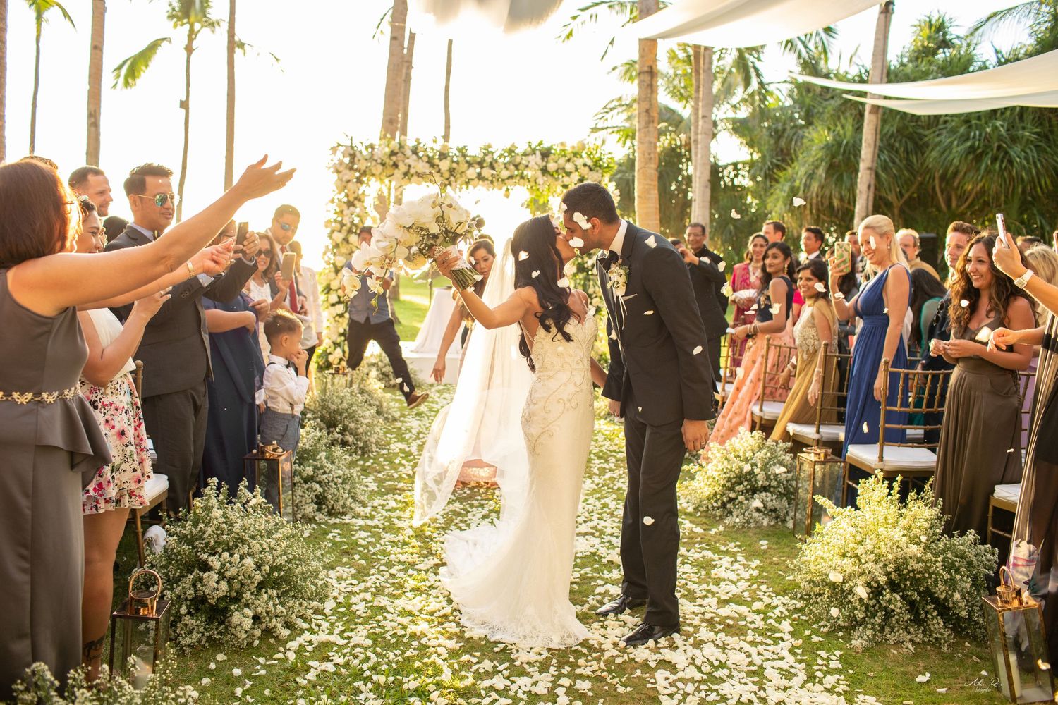 Bride and groom share a kiss while walking down a flower-lined outdoor wedding aisle surrounded by cheering guests.